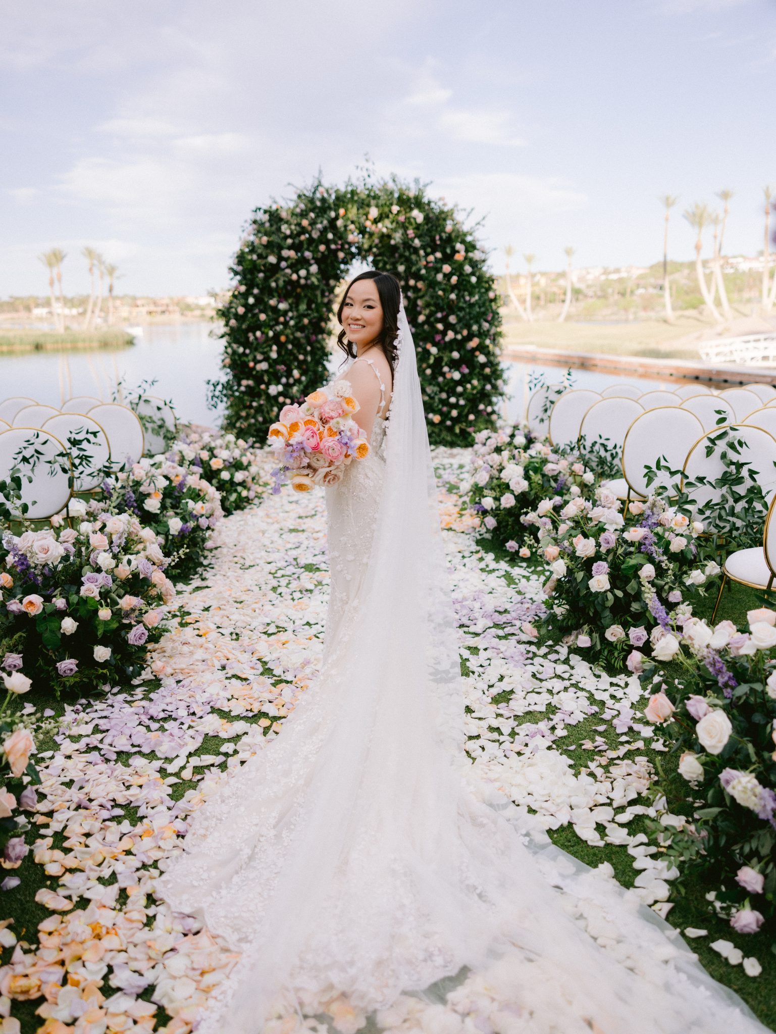 Bride posing with bouquet at a luxury Las Vegas wedding Rocky Mountain Bride – A stunning bride in a lace gown stands before a lush floral arch, holding a pastel bouquet, with a serene lake in the background at a luxury Las Vegas wedding Rocky Mountain Bride.