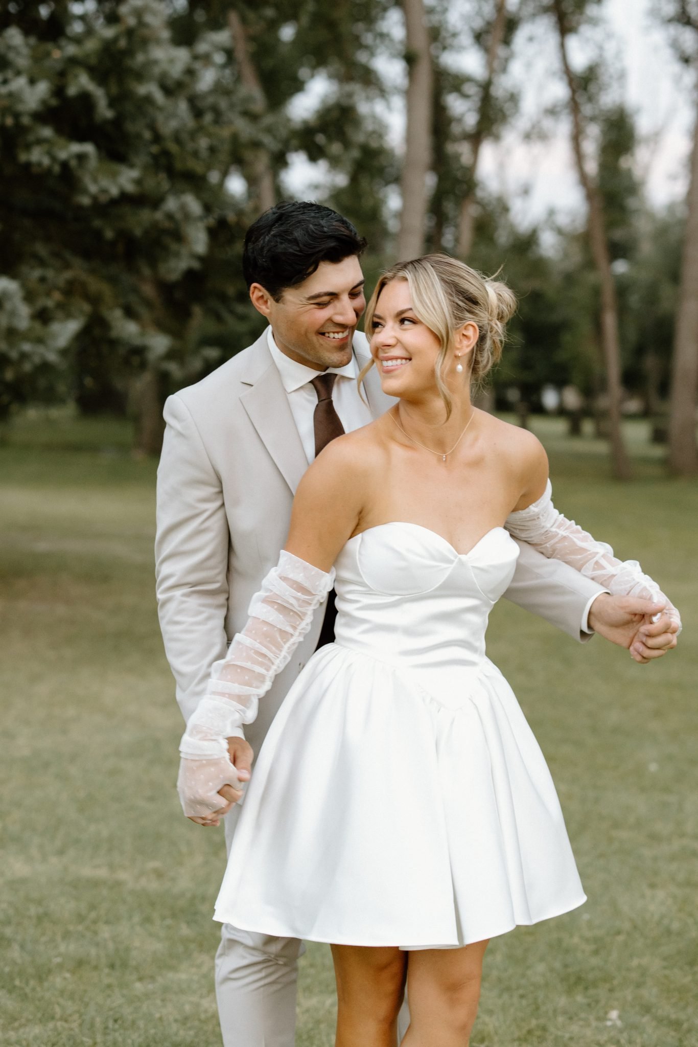 Bride in a short white dress with sheer sleeves and groom in a light suit smiling during an Outdoor Alberta wedding.