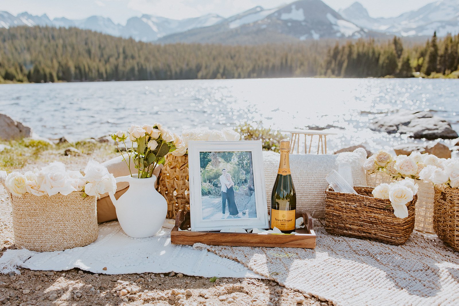 A picturesque lakeside proposal setup with white roses, framed photos, and champagne, set against a stunning mountain backdrop for a Brainard Lake Proposal.
