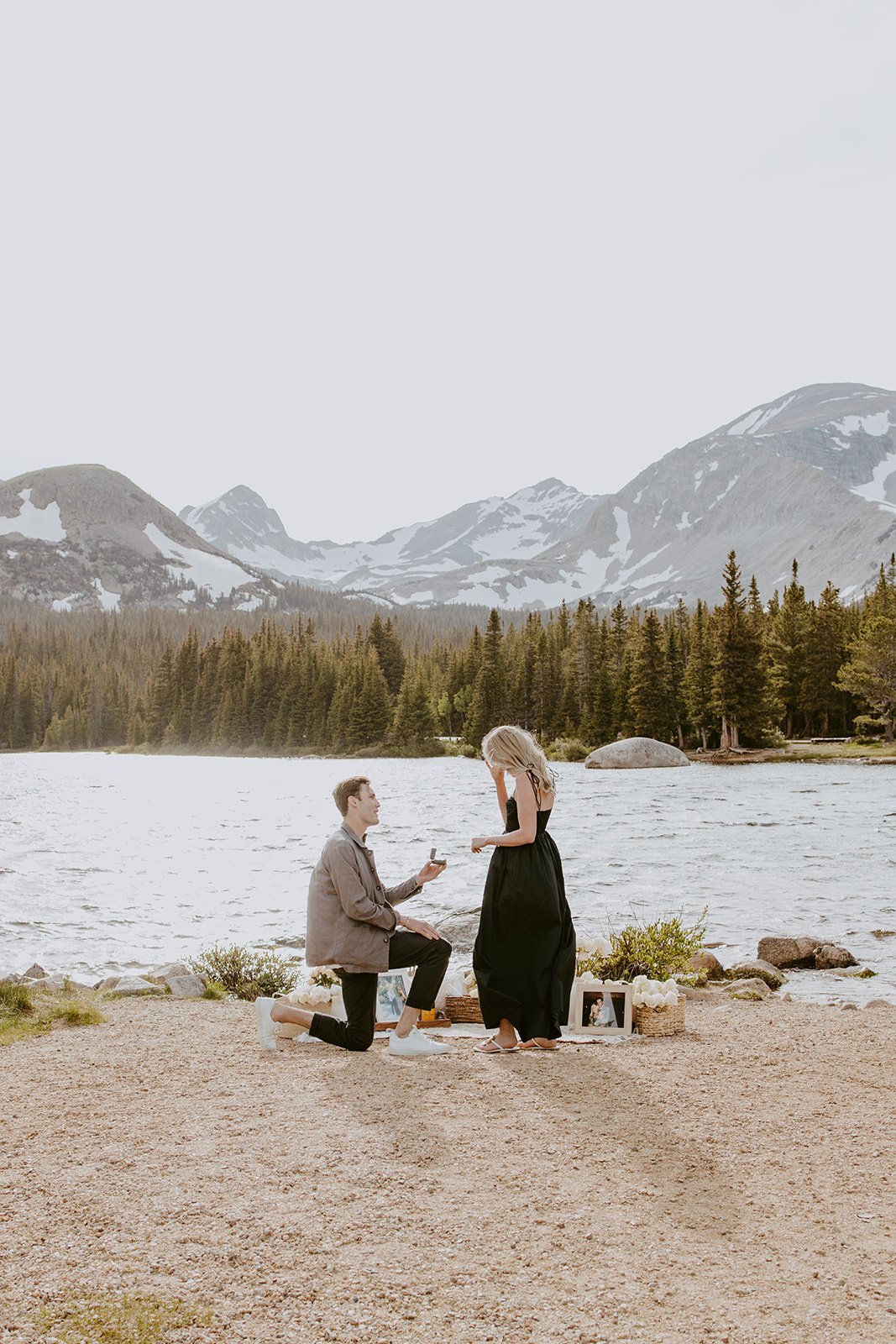 A groom-to-be kneels on one knee, proposing to his partner by the shimmering water with snow-capped peaks in the background at their Brainard Lake Proposal.
