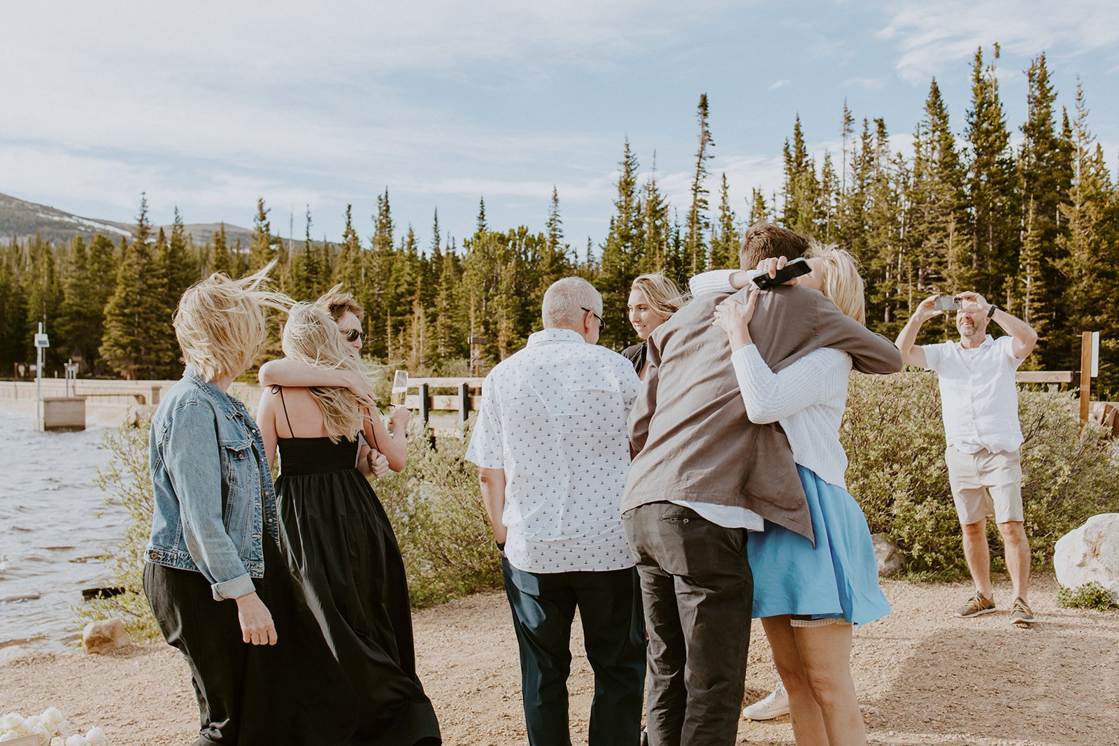 The newly engaged couple embraces their loved ones, celebrating their Brainard Lake Proposal with hugs and champagne by the water’s edge.