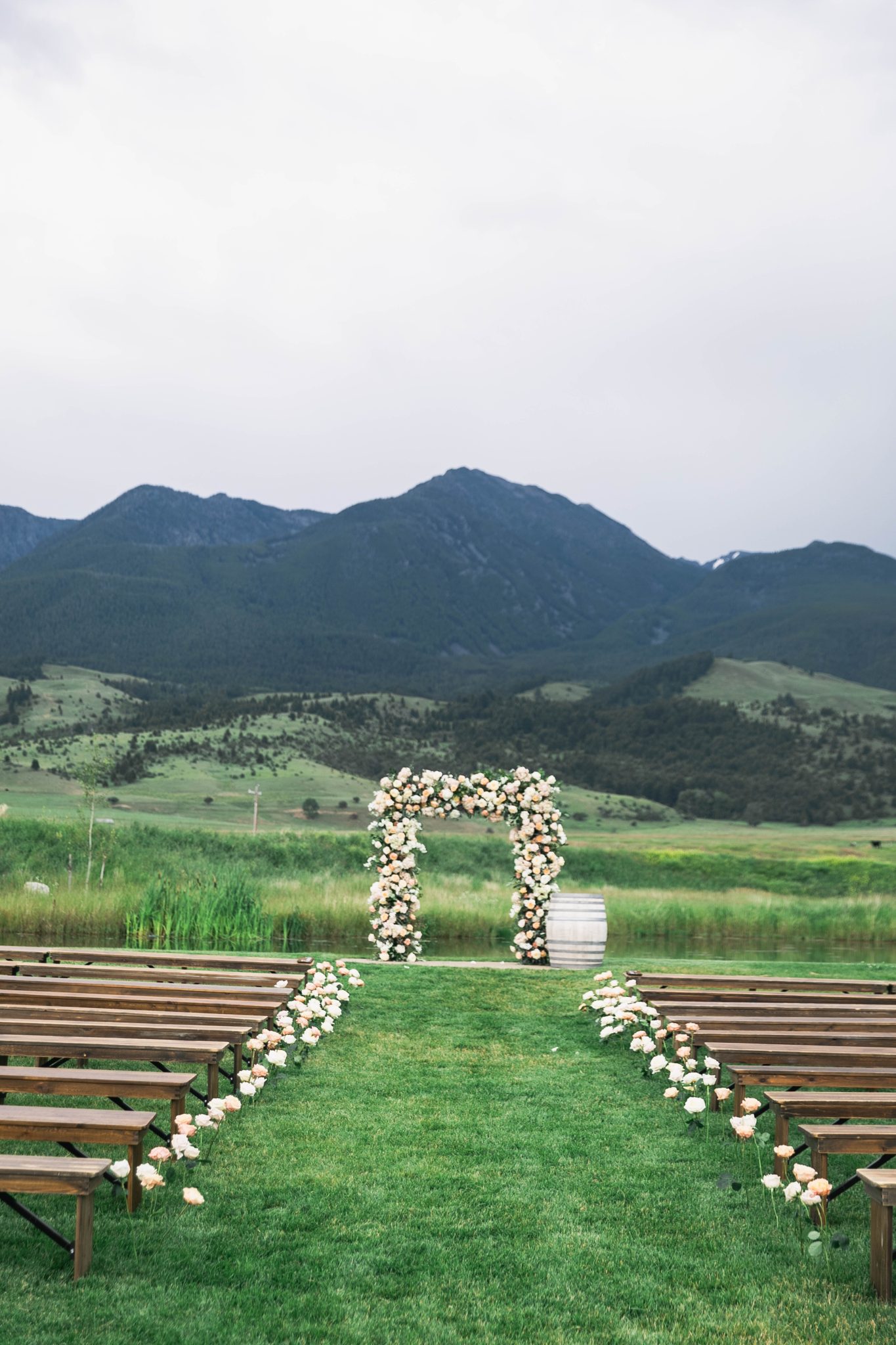 A breathtaking outdoor ceremony setup at a Copper Rose Ranch Montana wedding rocky mountain bride, featuring a floral-covered arch with soft blush and ivory roses against a stunning mountain backdrop.