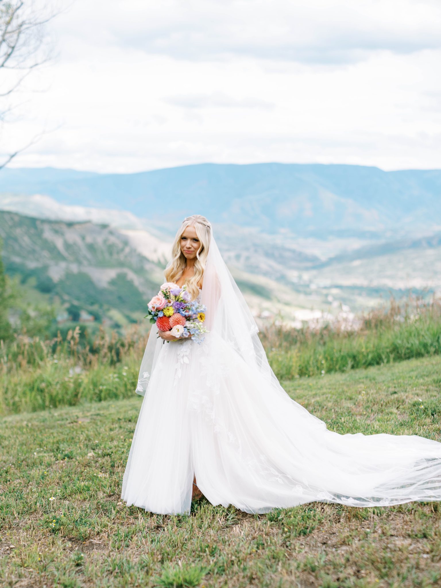 A stunning bride in a flowing tulle gown with floral appliqués poses against a breathtaking mountain backdrop at an Elegant Summer Wedding in Snowmass Colorado.