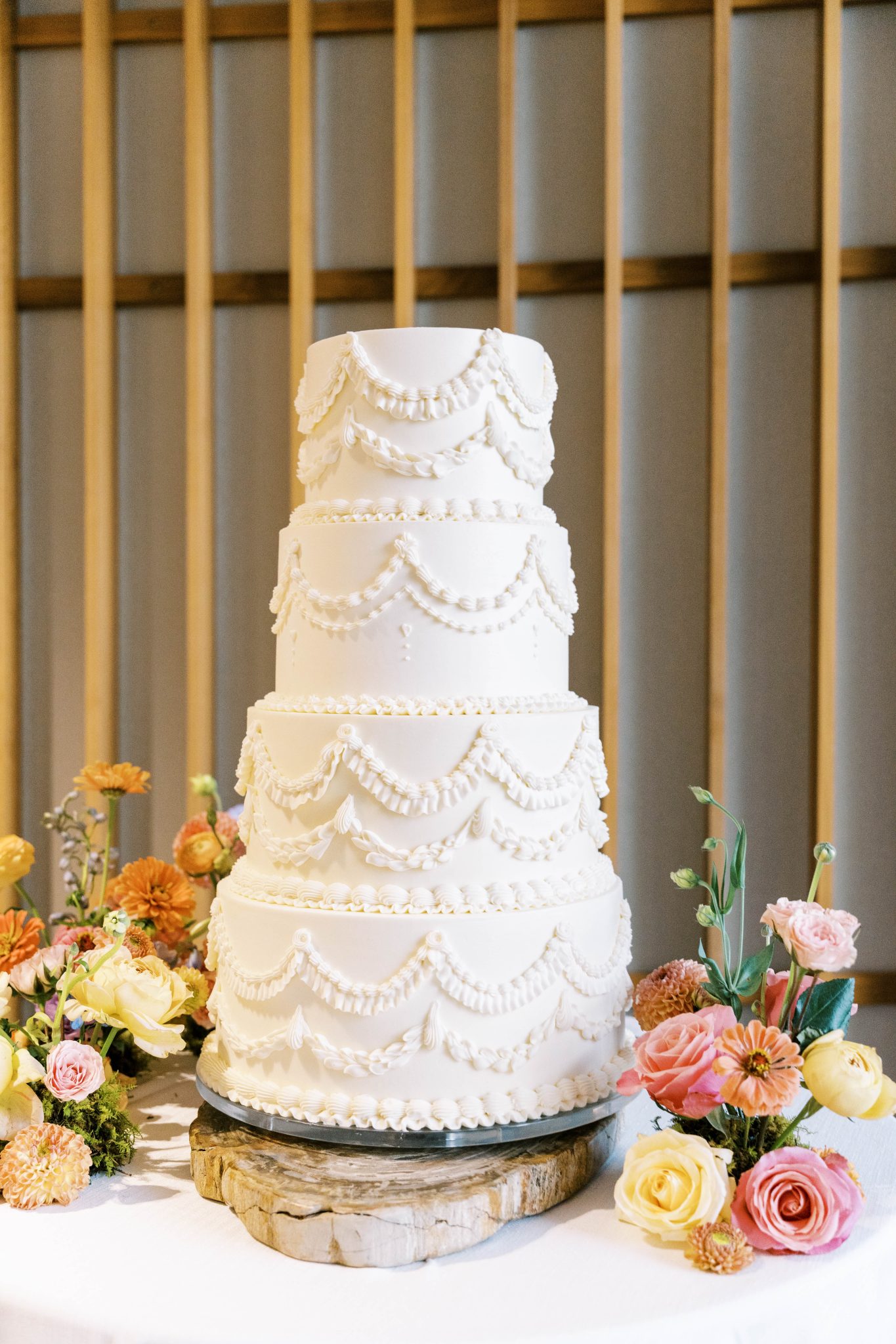 A luxurious four-tier white wedding cake with intricate piping and vintage-inspired details, surrounded by vibrant florals at an Elegant Summer Wedding in Snowmass Colorado.