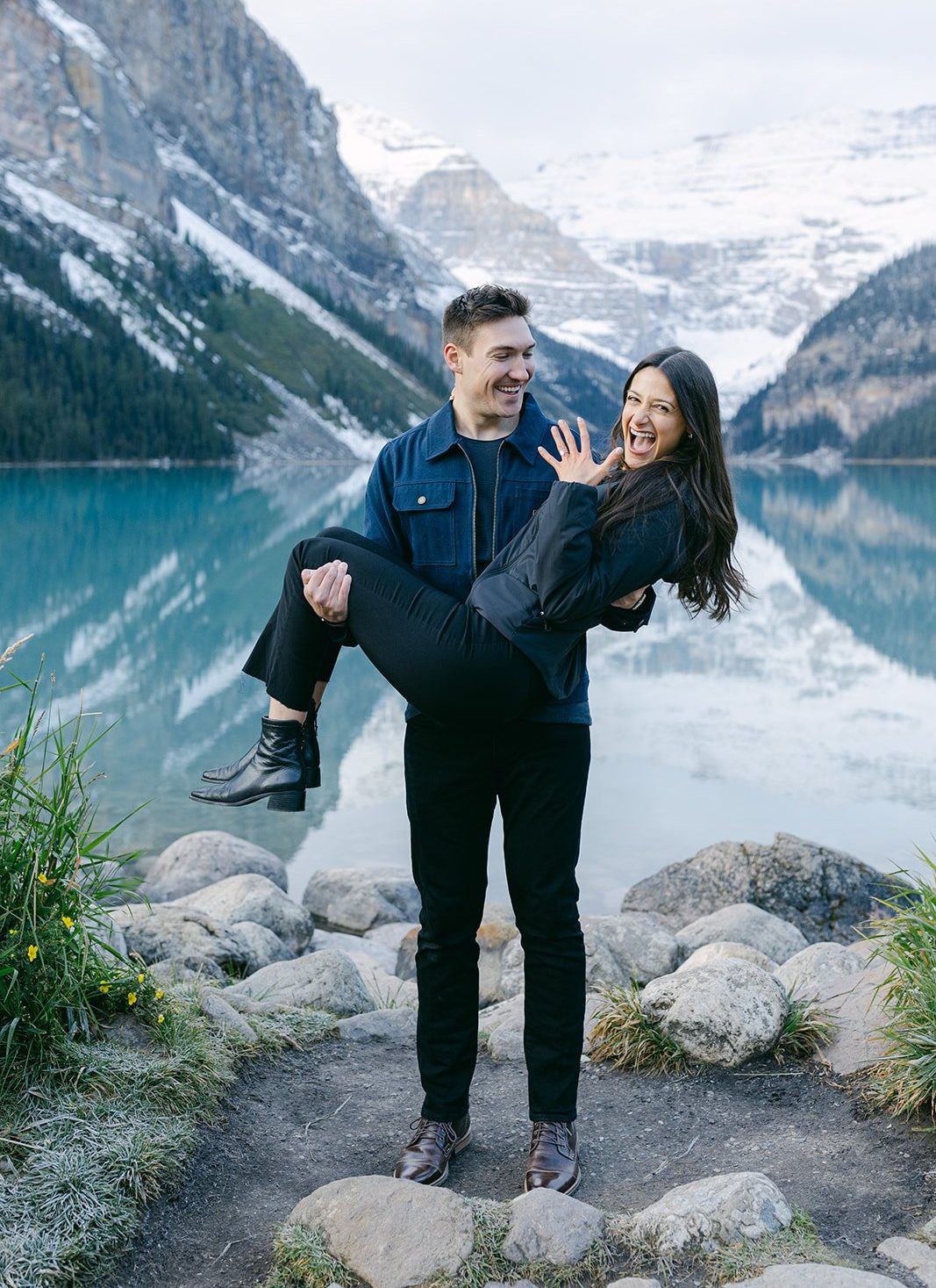 Lake Louise Banff National Park Surprise Proposal – A newly engaged couple shares a playful moment as the groom-to-be lifts his fiancée, both laughing in front of the breathtaking Lake Louise landscape.