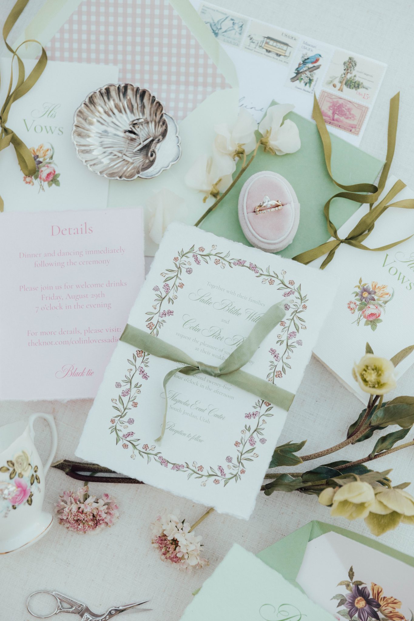 Flat lay of delicate wedding invitations with floral details and sage green accents at Magnolia Event Center Wedding Inspiration Rocky Mountain Bride, styled with vintage stamps, a velvet ring box, and silk ribbon.