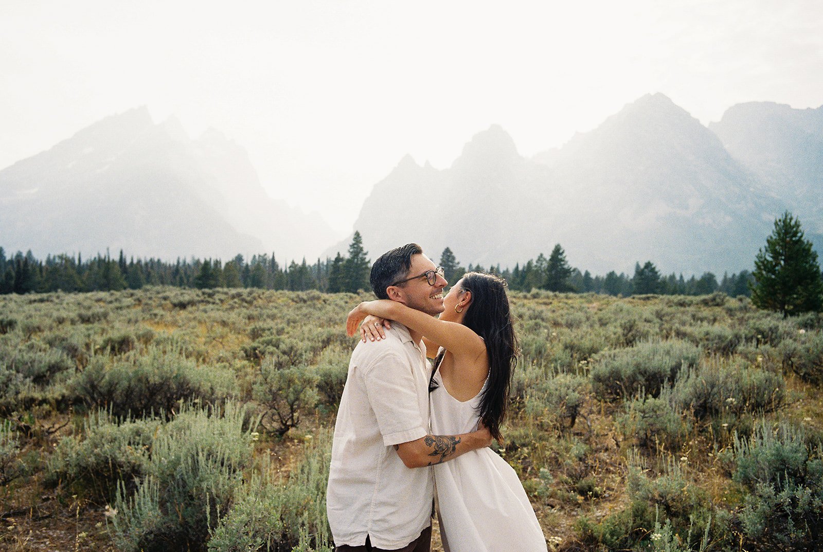 A loving couple shares a quiet moment in a sunlit forest, dressed in soft neutral tones for their National Park Day Before Wedding Session.