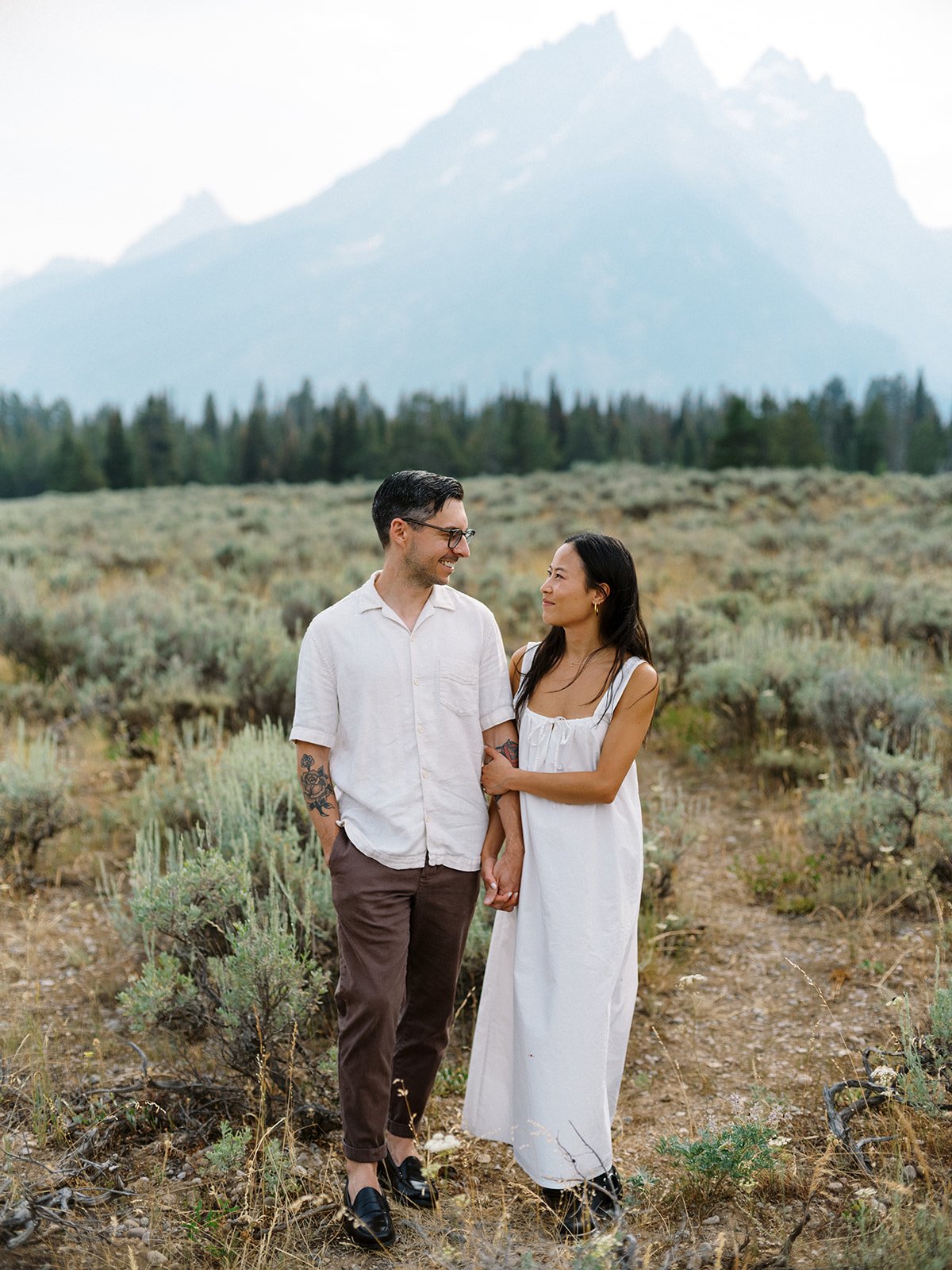 A couple walks hand in hand through a scenic meadow with towering mountain peaks in the background during their National Park Day Before Wedding Session.
