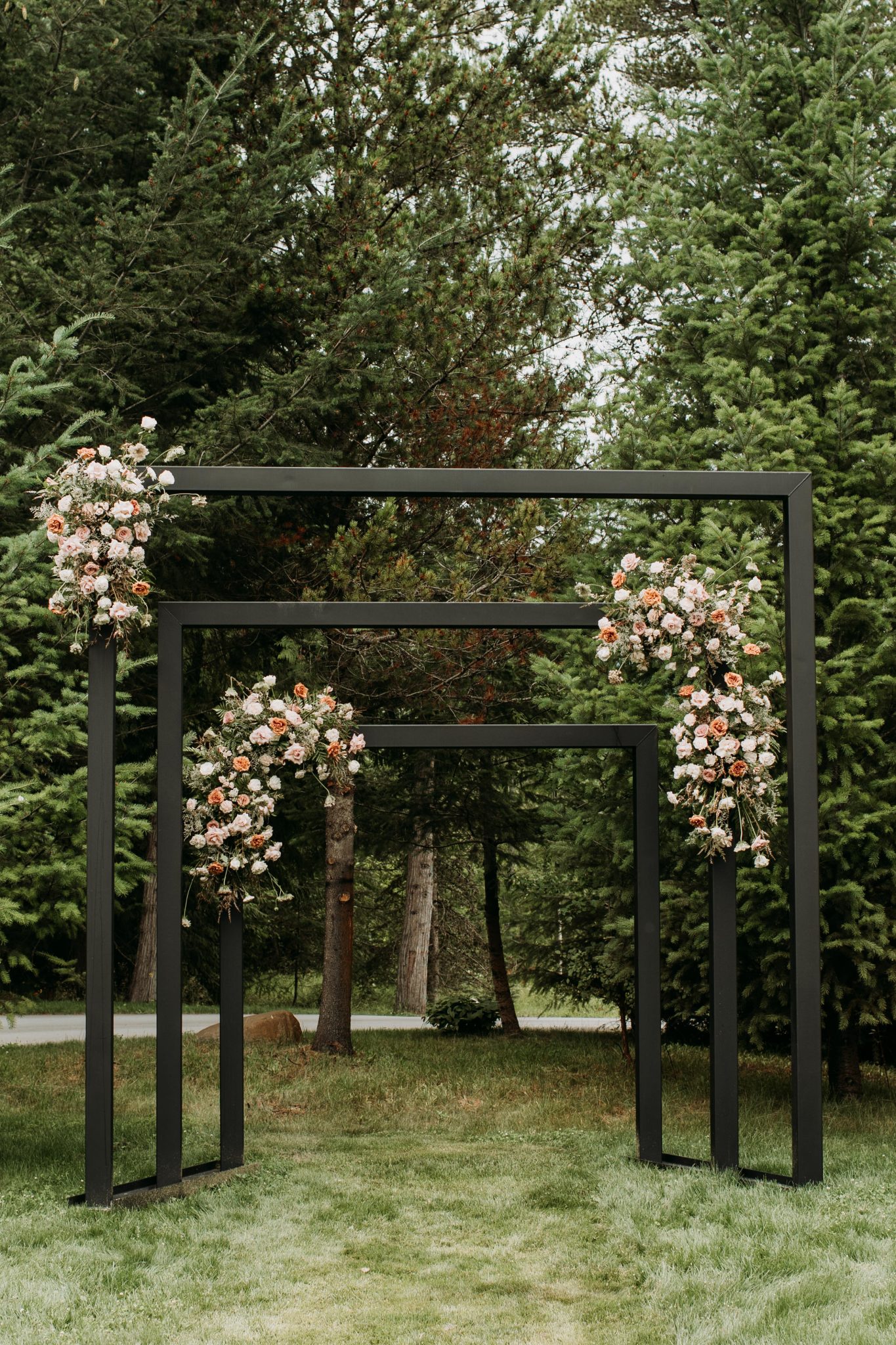 A modern black geometric ceremony arch adorned with blush and peach florals stands against lush greenery, creating a striking altar for an Outside summer wedding in BC rocky mountain bride.