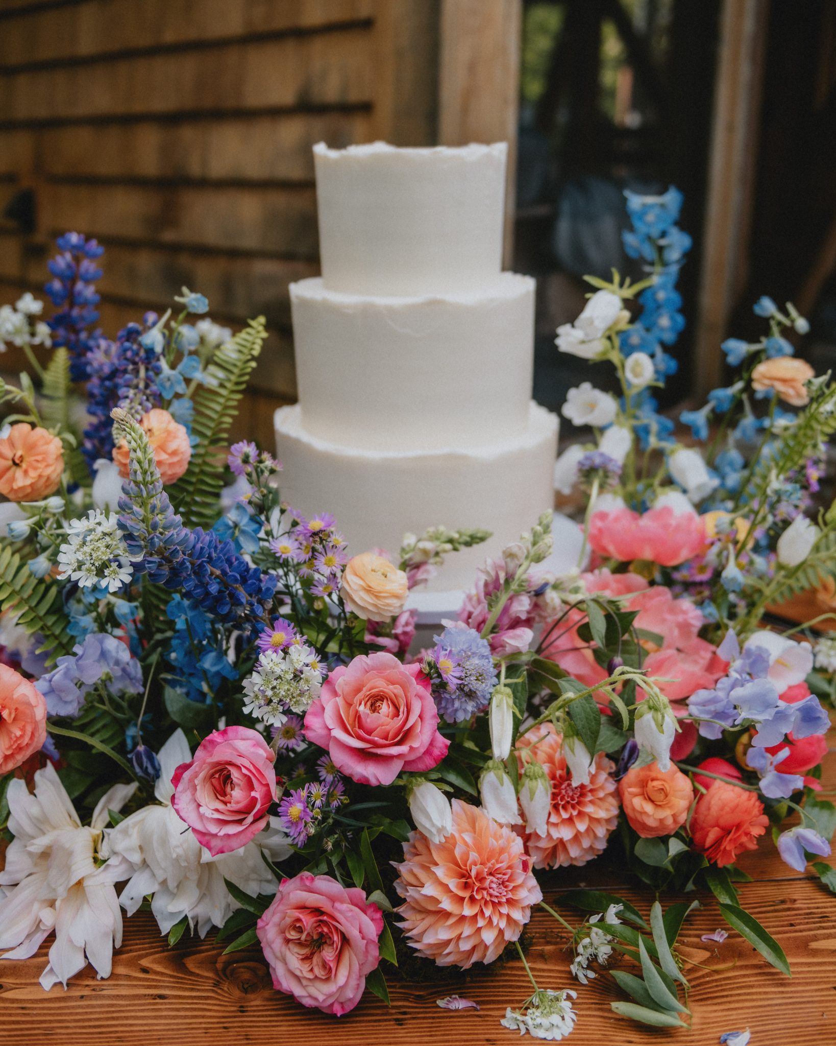 A stunning three-tier white wedding cake surrounded by bold pink, orange, and blue florals, adding a colorful touch to a Vibrant Montana outside wedding.