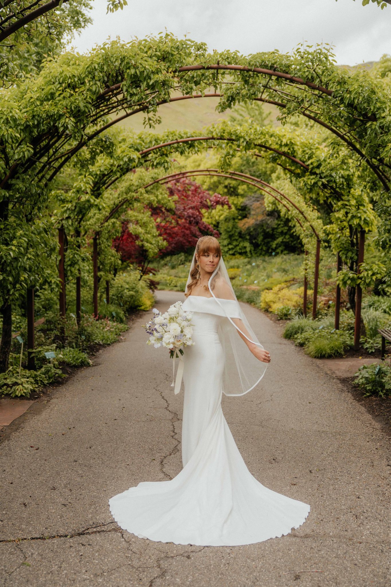 A bride in an elegant off-the-shoulder gown with a long veil, standing under a lush green archway at a Spring Salt Lake City Wedding.