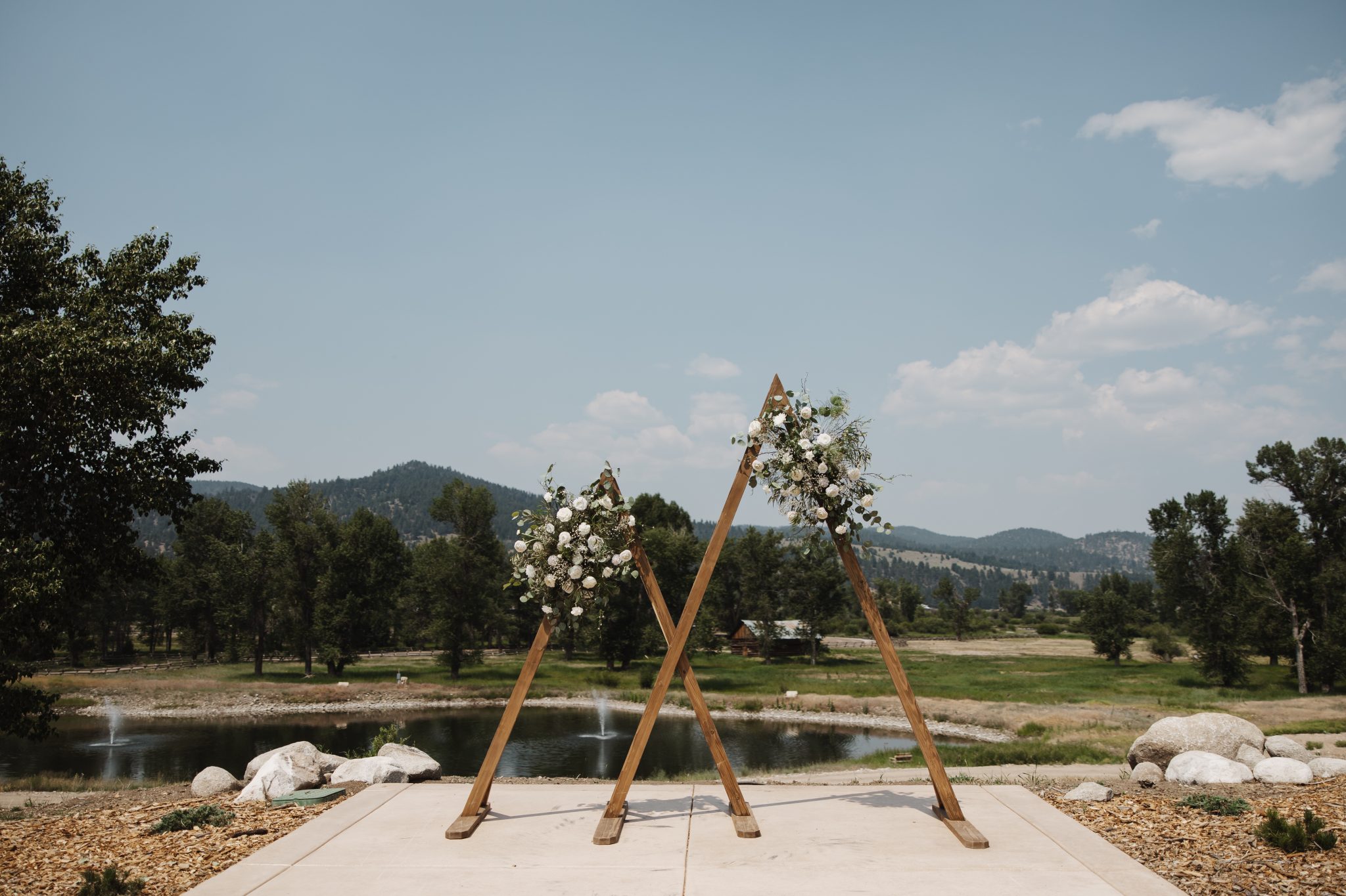 A rustic wooden triangular arch adorned with white florals and greenery stands against a picturesque mountain view at The Meadows on Rock Creek White Wedding.