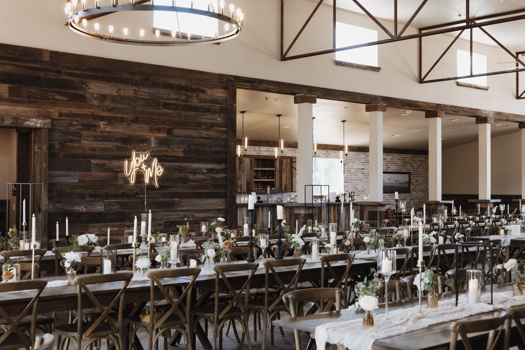 A warm and elegant reception space featuring wooden accents, candlelit tables, and a romantic “You + Me” neon sign at The Meadows on Rock Creek White Wedding.