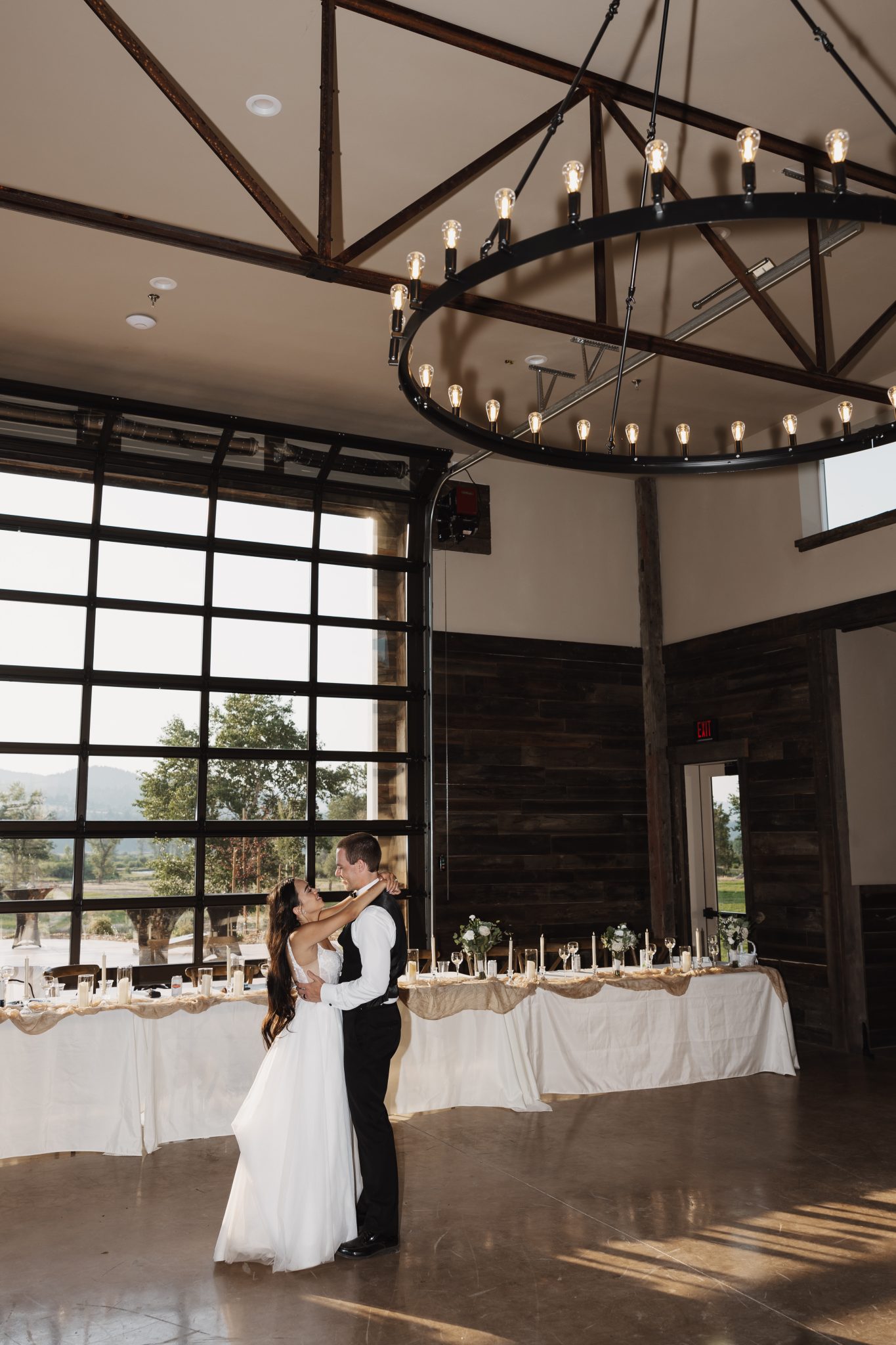 The newlyweds share a romantic first dance under a statement chandelier, surrounded by candlelit tables at The Meadows on Rock Creek White Wedding.