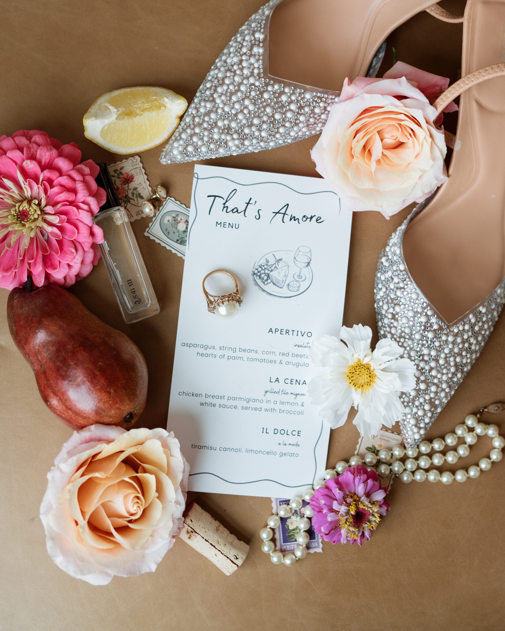 Flat lay of a White Barn wedding inspiration featuring pearl-embellished heels, floral details, a vintage-inspired menu card, and a romantic ring with a pearl accent.