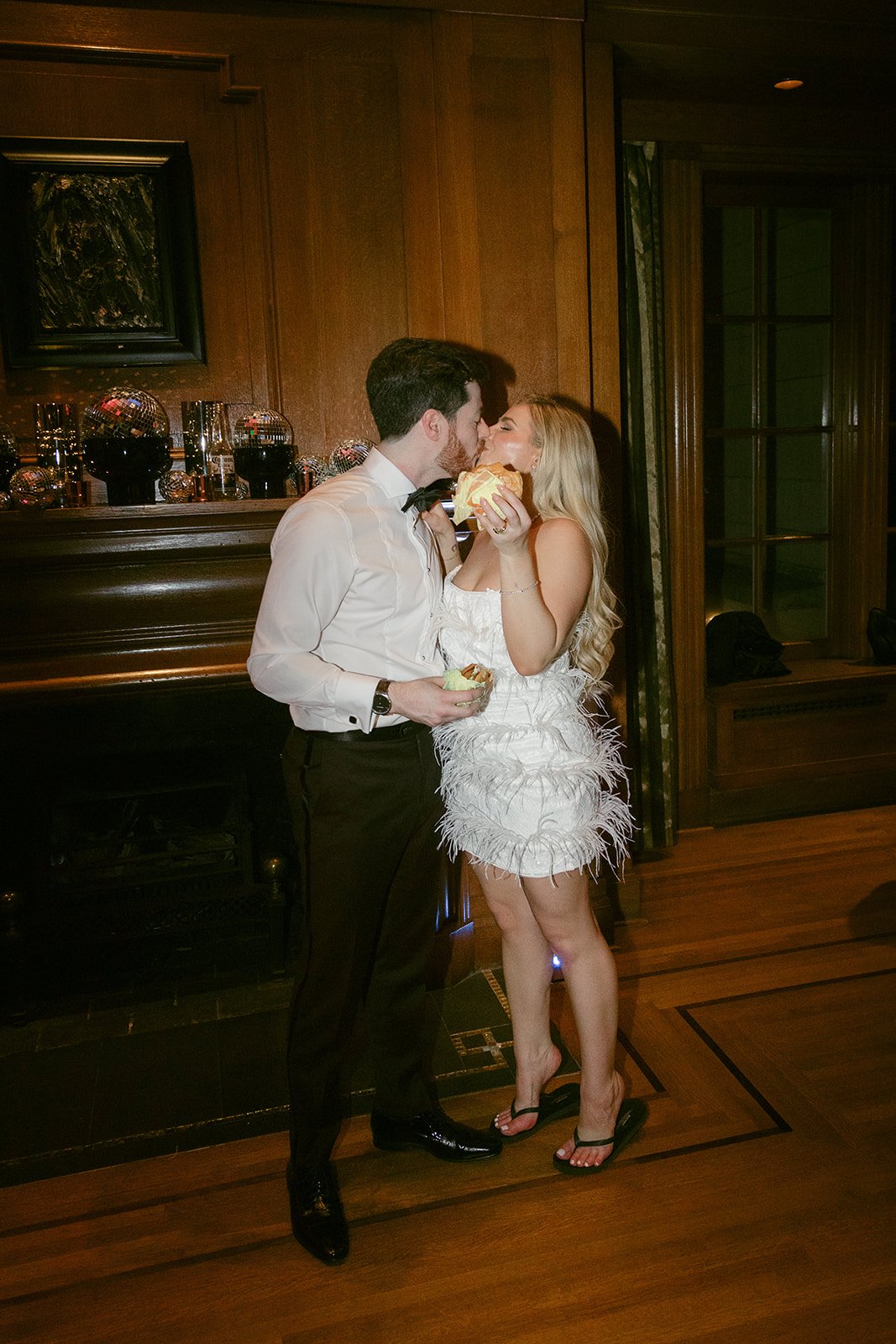Bride and groom enjoy a fun late-night snack at their elegant Vancouver wedding Rocky Mountain Bride, sharing a kiss while holding burgers in a cozy reception lounge with chic décor.