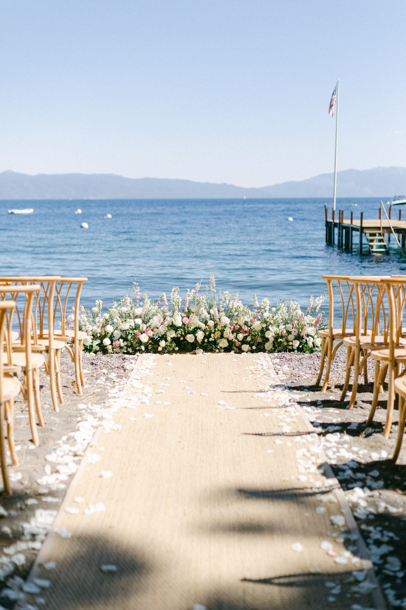 A breathtaking lakeside wedding ceremony setup at lake tahoe wedding rocky mountain bride, featuring wooden chairs, a petal-lined aisle, and lush floral arrangements against the backdrop of crystal blue waters and mountain views.