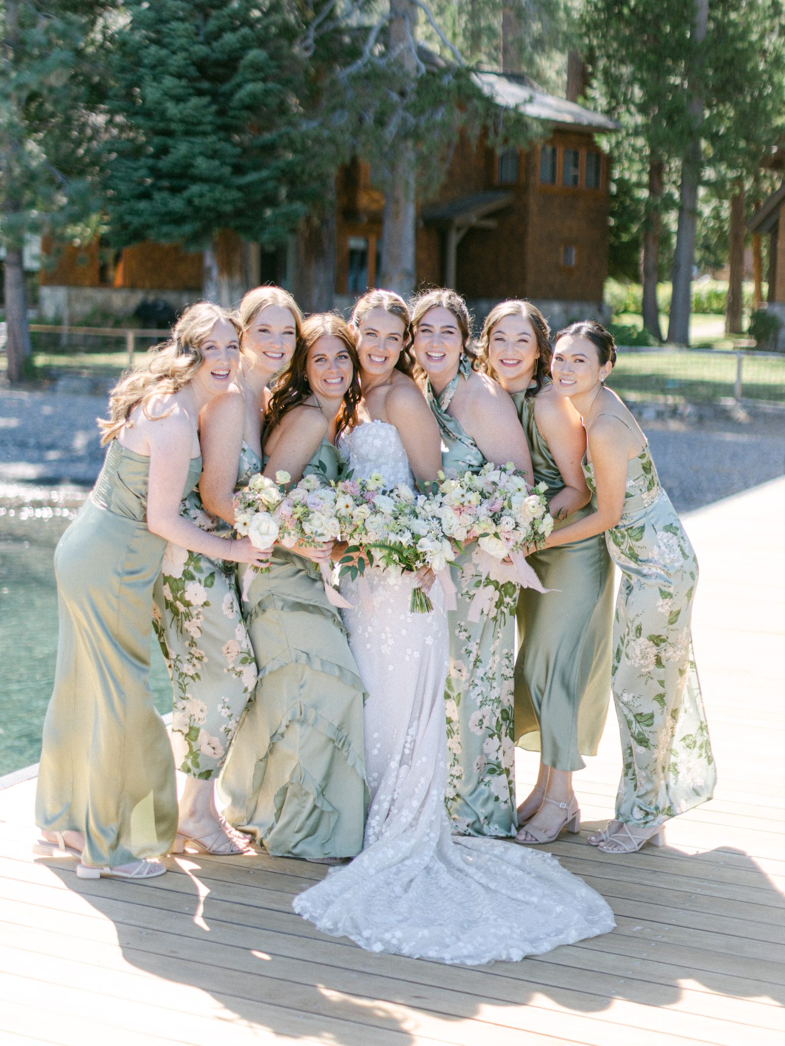 The bride and her bridesmaids pose together on a wooden dock in elegant sage green and floral dresses, holding pastel bouquets at a stunning lake tahoe wedding rocky mountain bride celebration.