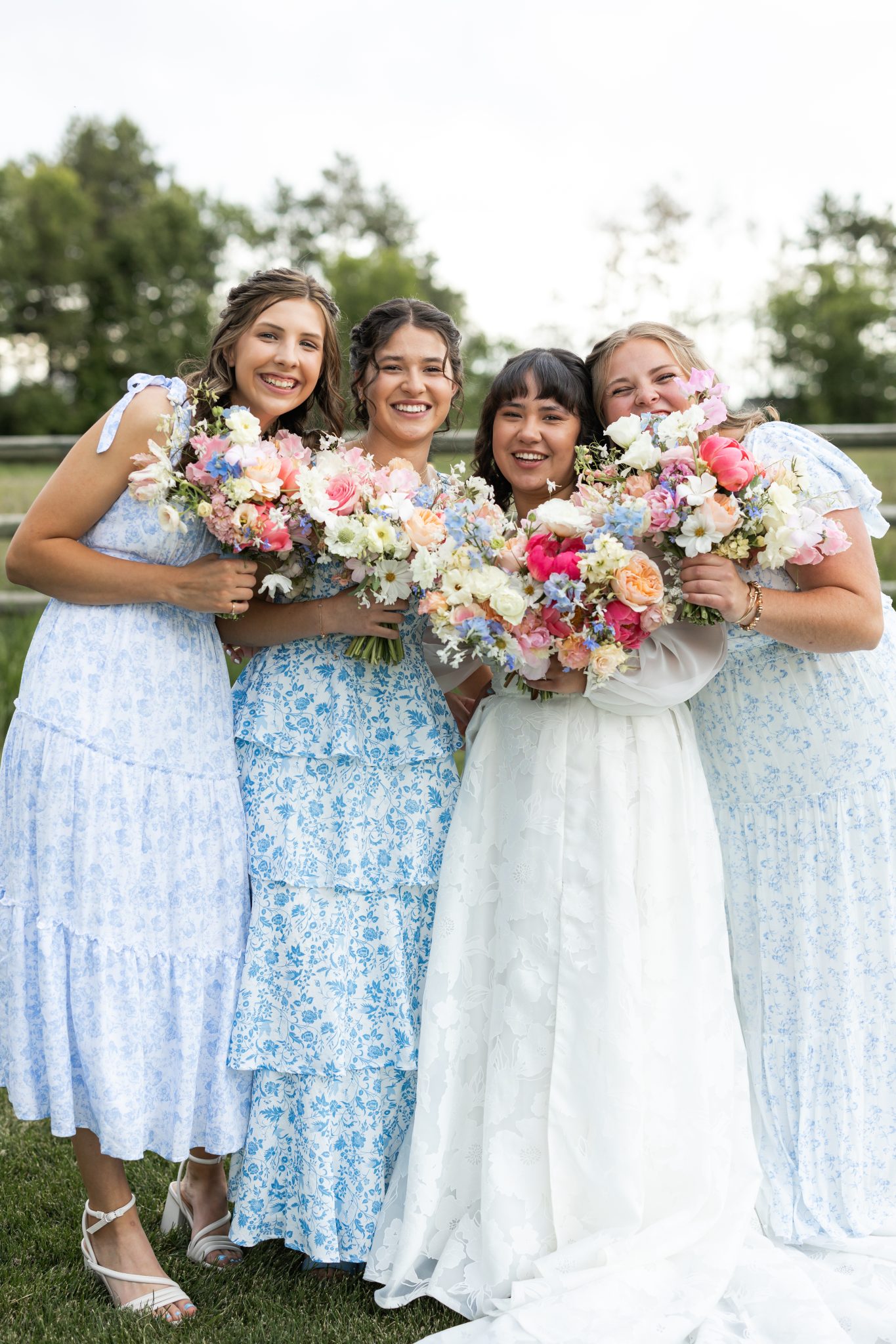 The bride in a floral-textured white wedding dress, posing with her bridesmaids in blue floral dresses, all holding vibrant bouquets with pink, white, and blue flowers. A joyful moment captured by Buckley Photography spring Idaho weding rocky mountain bride.