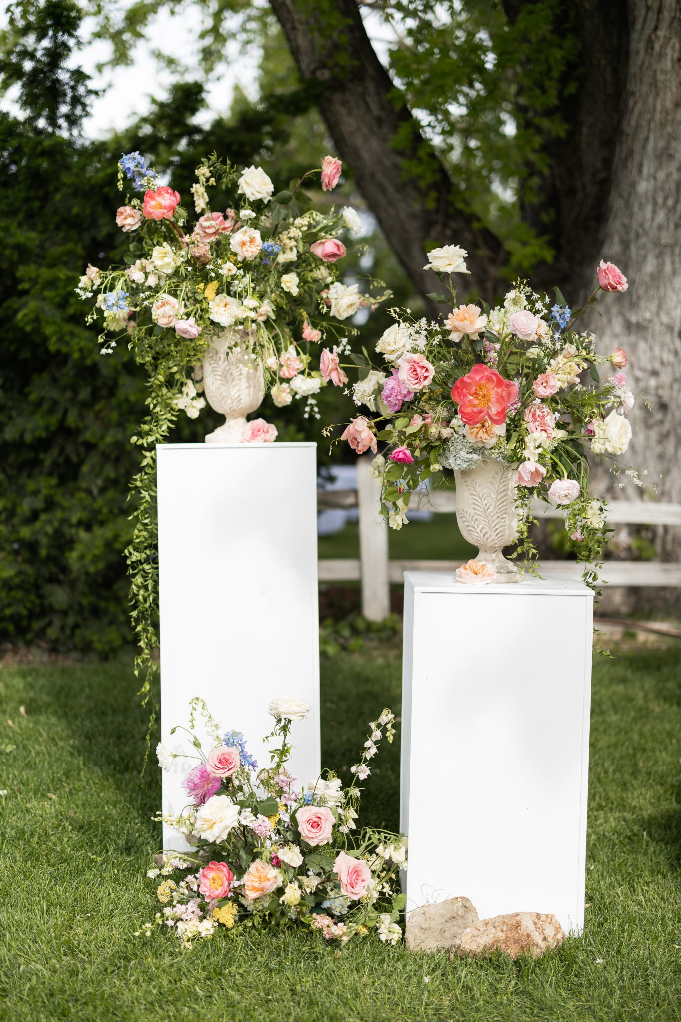 Stunning floral displays with pink, blue, and peach-toned blooms set on white pedestals, creating a dreamy outdoor ceremony setting. Captured by Buckley Photography spring Idaho wedding rocky mountain bride.