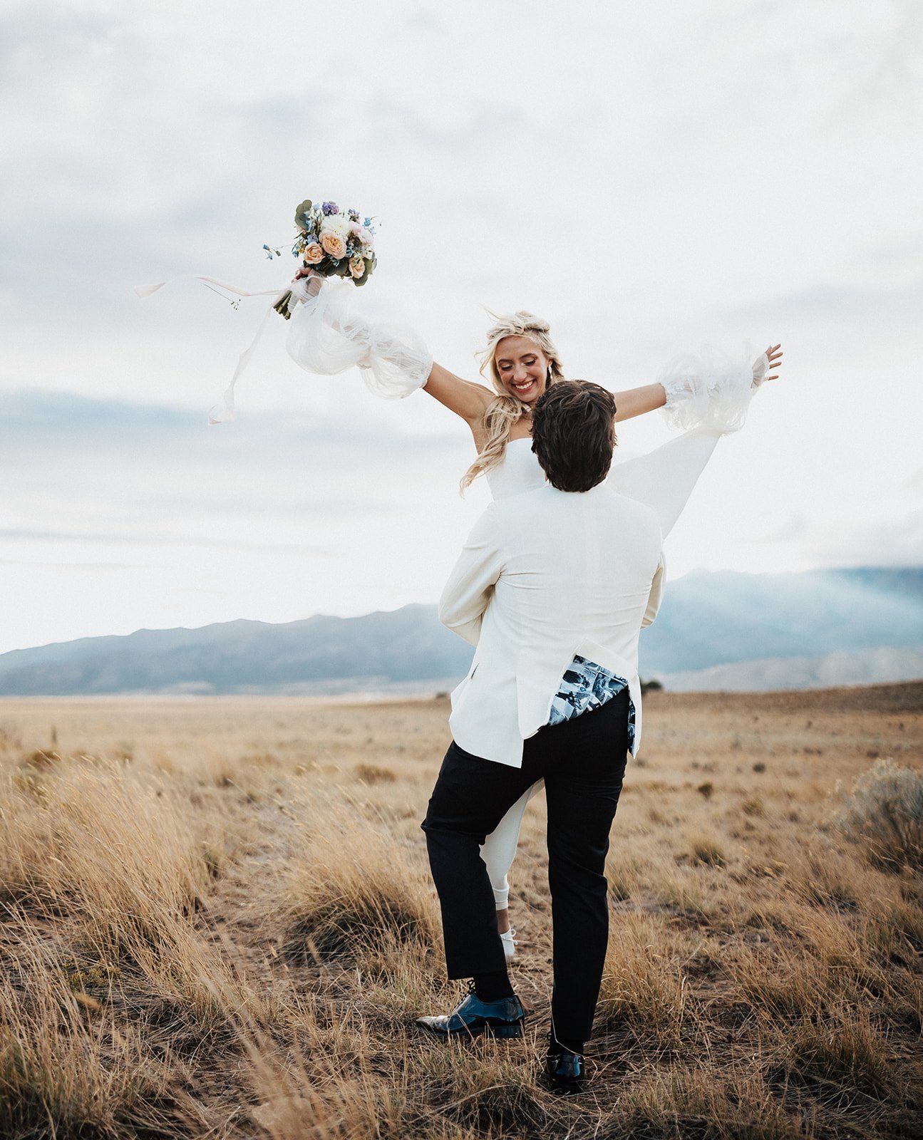 Joyful fall Utah wedding photo of the bride holding her bouquet while being lifted by the groom in a golden grass field.