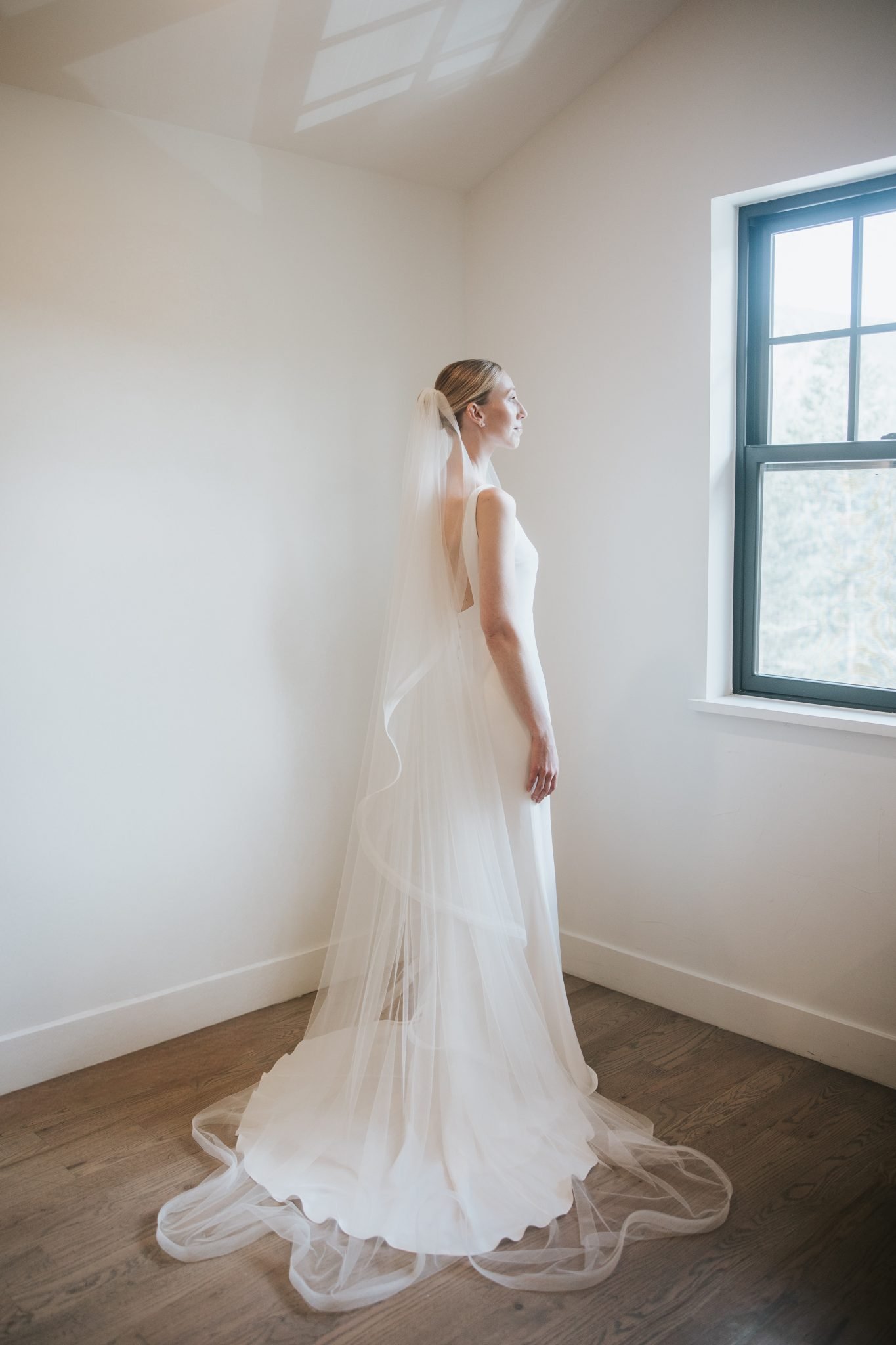 A bride in a sleek, modern wedding gown with a flowing veil stands by a window, capturing the elegance of a North Start Gathering wedding in Idaho Springs Rocky Mountain Bride.
