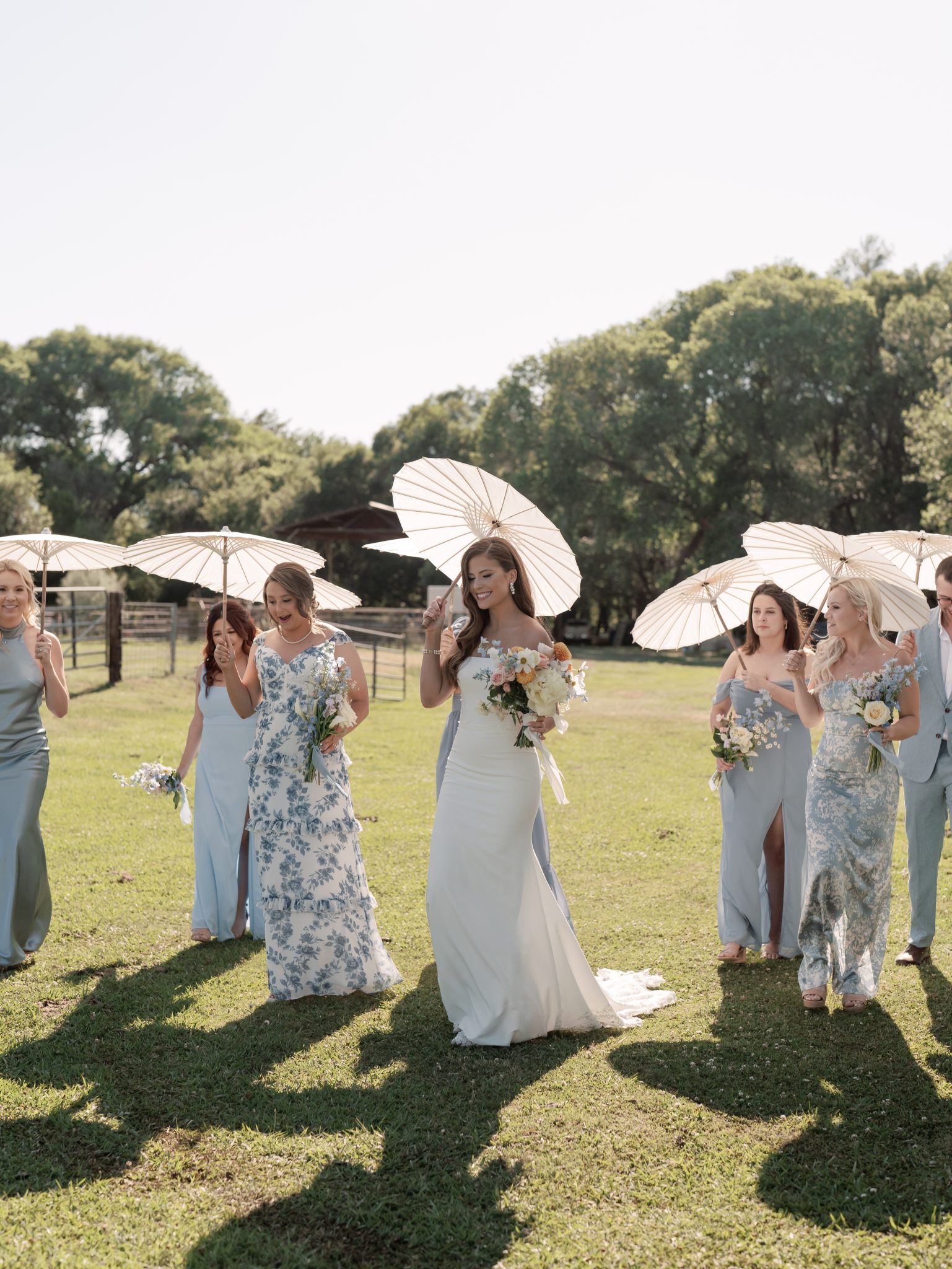 Bridal party in mismatched blue dresses holding white parasols on a sunny day at a Private Property Saje Photography Arizona Wedding.