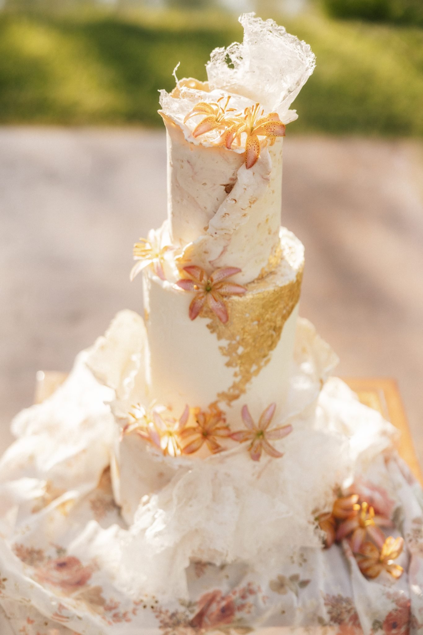 Artistic wedding cake adorned with delicate sugar flowers and gold accents, styled against a soft floral fabric, showcasing Summer montana wedding inspiration rocky mountain bride.
