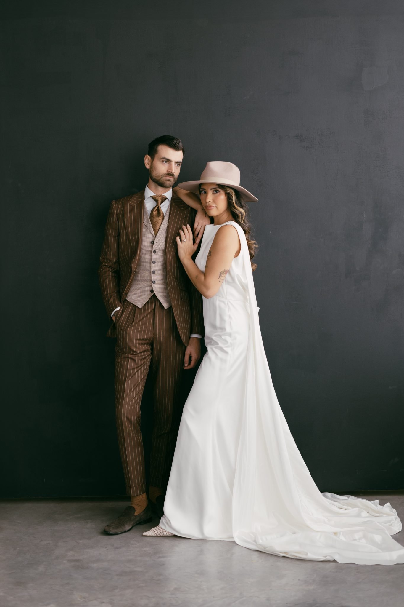 Fashion-forward bride in a sleek white gown and wide-brim hat posing with groom in a brown pinstripe suit for The Gallery Studio wedding inspiration rocky mountain bride.