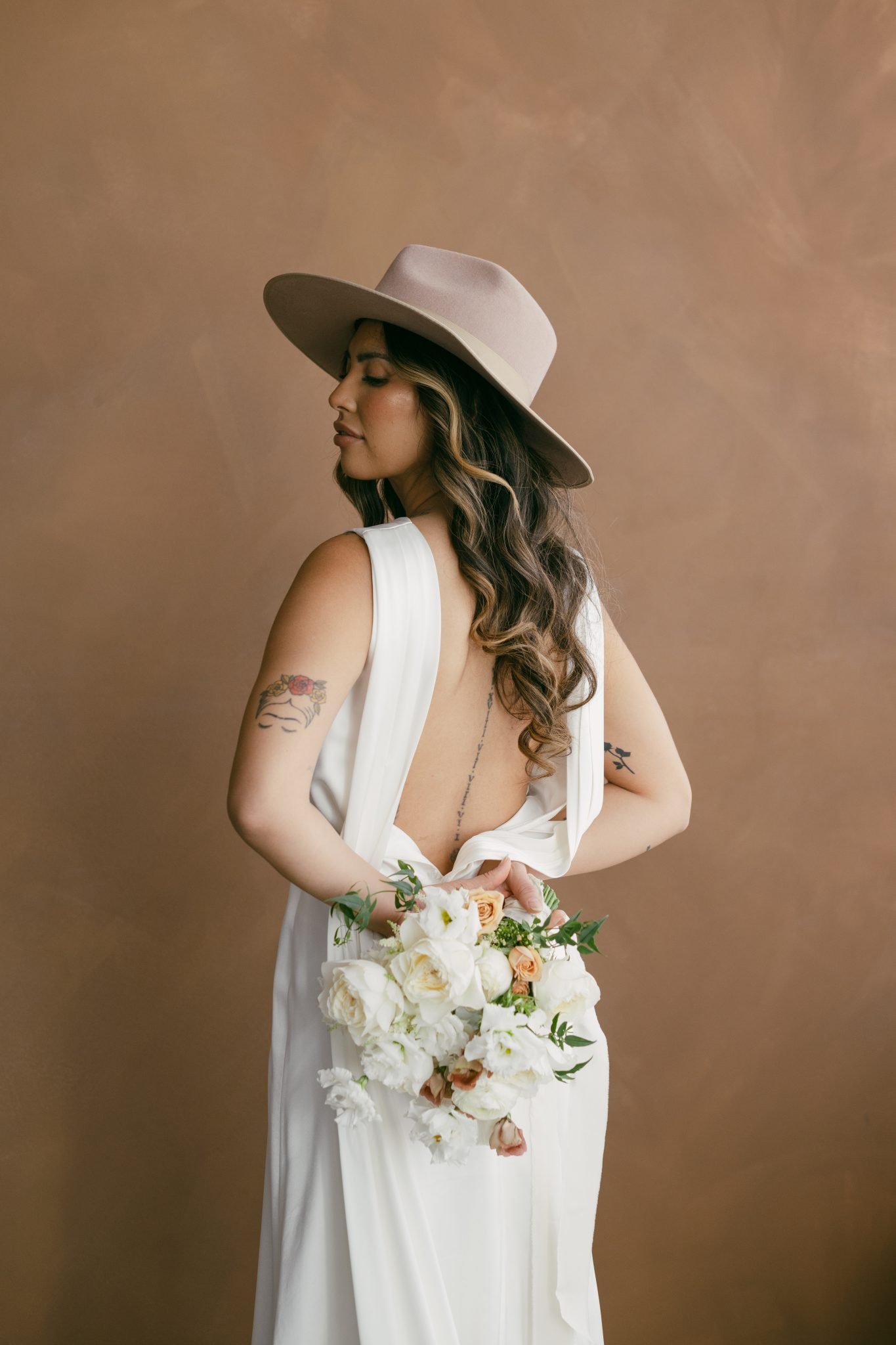 Bride in a backless white gown and taupe hat holding a white and blush floral bouquet behind her for The Gallery Studio wedding inspiration rocky mountain bride.