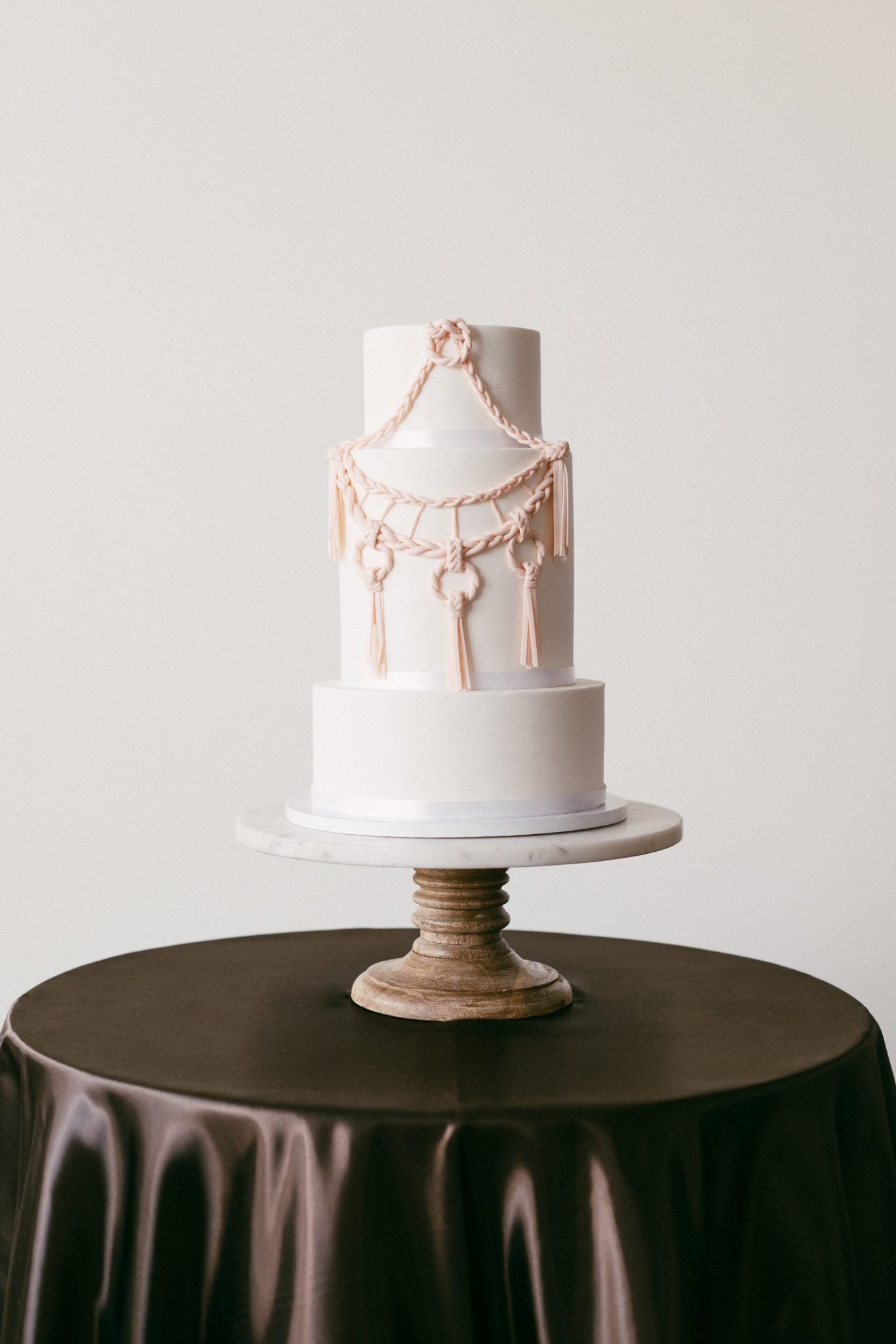 Three-tier white wedding cake with blush macramé detailing on a marble stand for The Gallery Studio wedding inspiration rocky mountain bride.