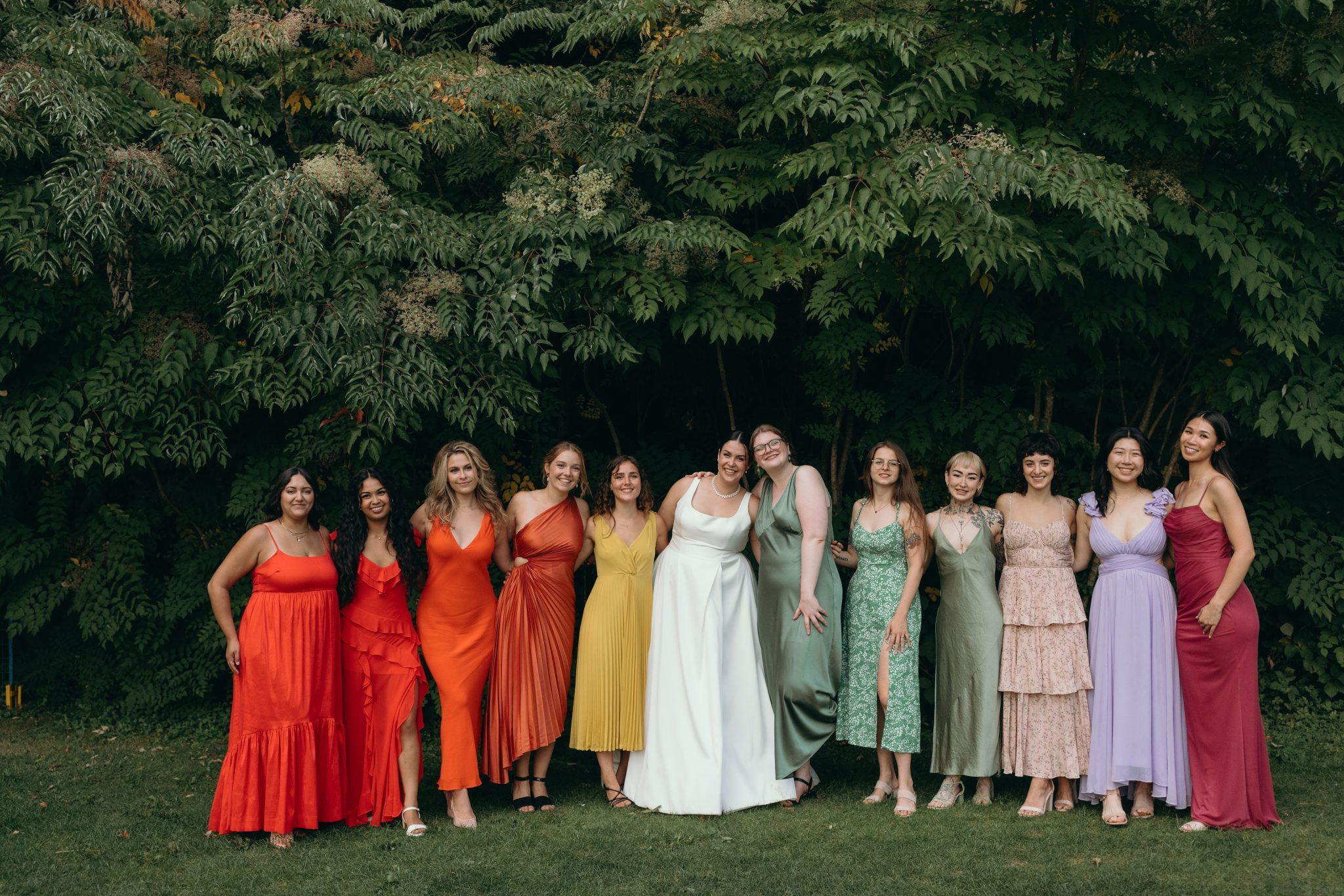 Group photo of the bride with bridesmaids in colorful dresses standing against a lush green backdrop at a Vibrant British Columbia Tented Wedding
