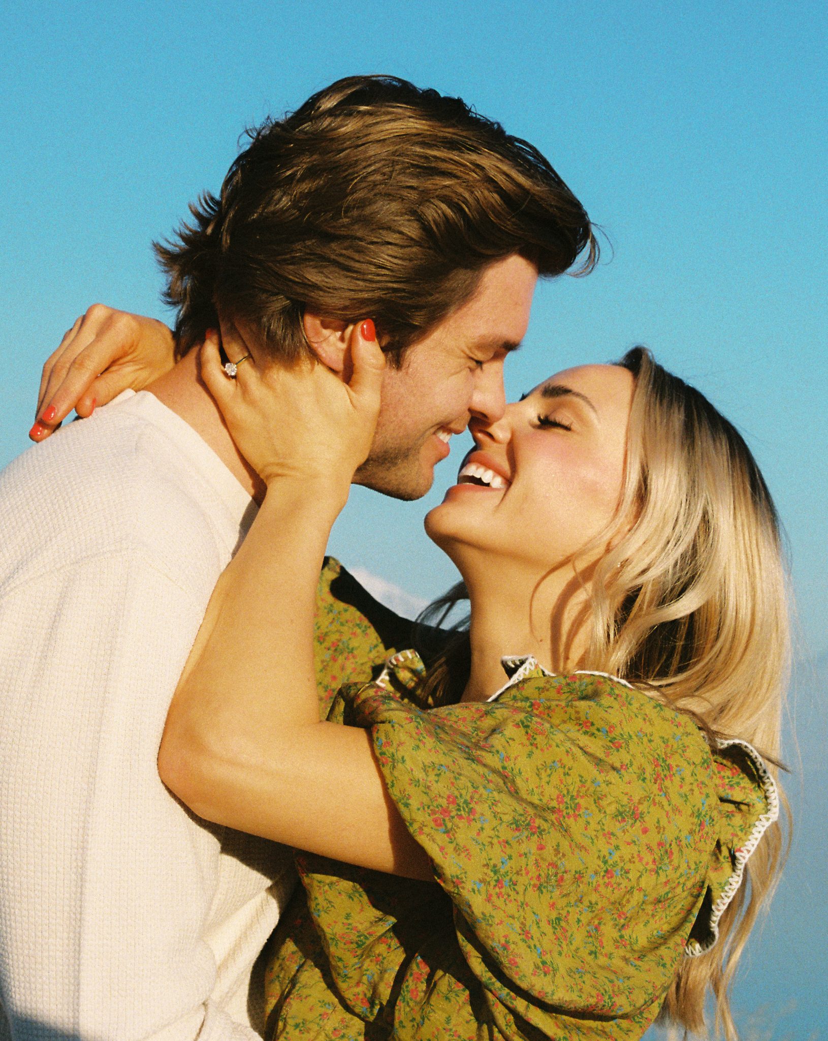 The couple embraces with noses touching, bathed in golden sunlight, as the bride-to-be wears a vintage-inspired green floral dress. A heartfelt moment from a Winter utah engagement session with Rachel Blank rocky mountain bride.