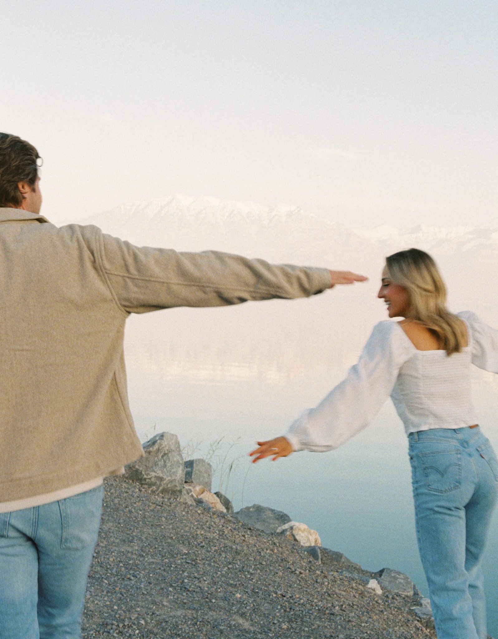A romantic shot of the couple holding hands and twirling near a winter lake, with soft pastel skies and snow-covered mountains in the background. A beautiful capture from a Winter utah engagement session with Rachel Blank rocky mountain bride.