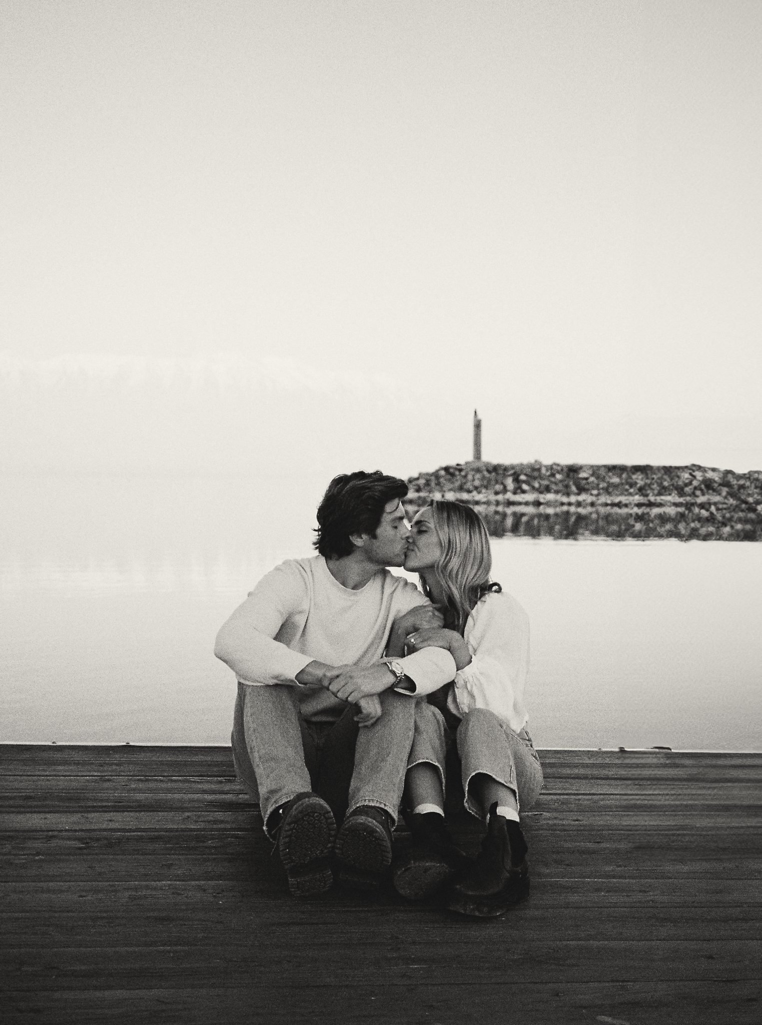 A couple shares an intimate kiss while sitting on a wooden dock, with snow-capped mountains and a calm lake in the background. Captured during a Winter utah engagement session with Rachel Blank rocky mountain bride.
