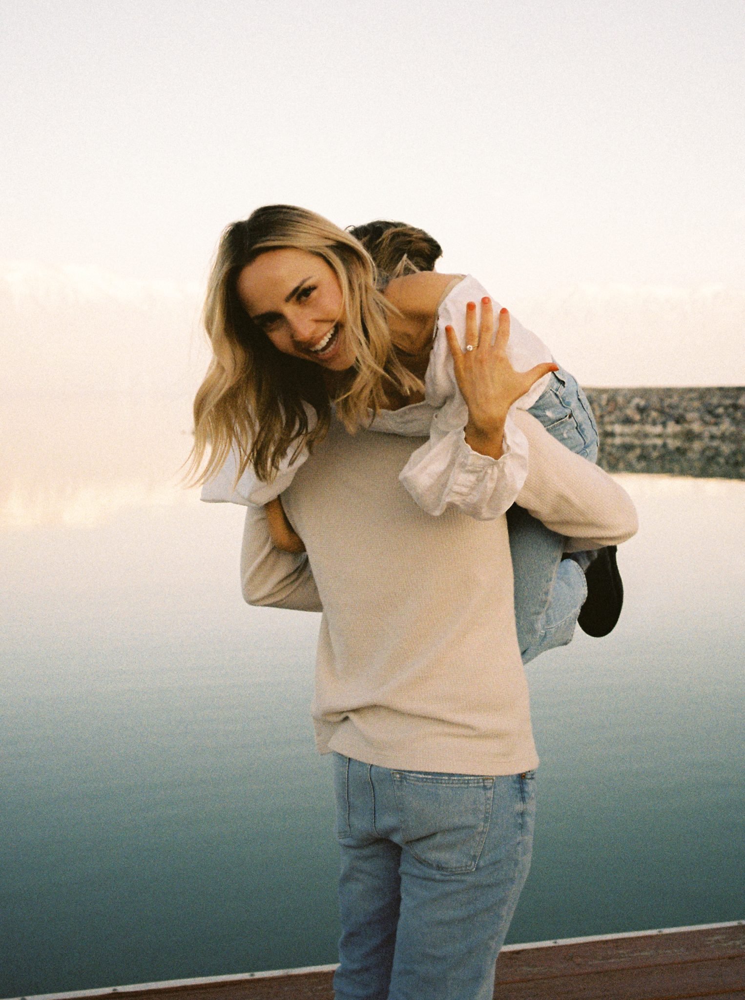 The bride-to-be playfully gets a piggyback ride from her fiancé, smiling and flashing her engagement ring against a serene winter lake backdrop. A candid shot from a Winter utah engagement session with Rachel Blank rocky mountain bride.