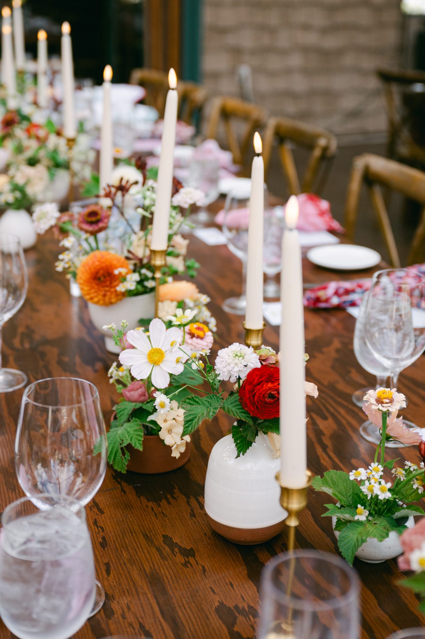 A wooden dinner table decorated with delicate floral arrangements in ceramic vases, tall taper candles, and rustic-chic tableware for an intimate wedding reception.