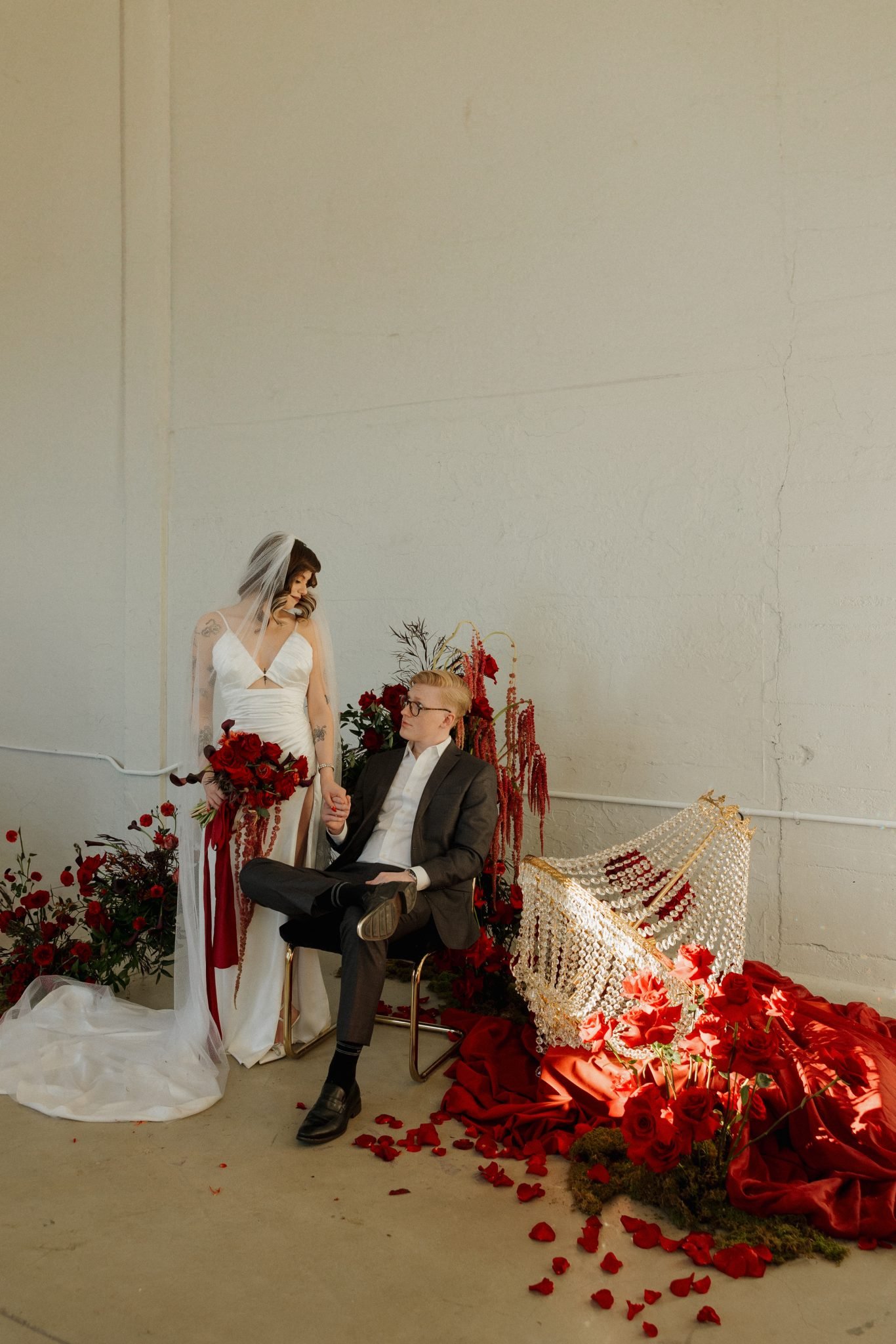 Stylish couple poses in front of moody red floral arch and vintage chandelier for moody red wedding inspiration rocky mountain bride feature.