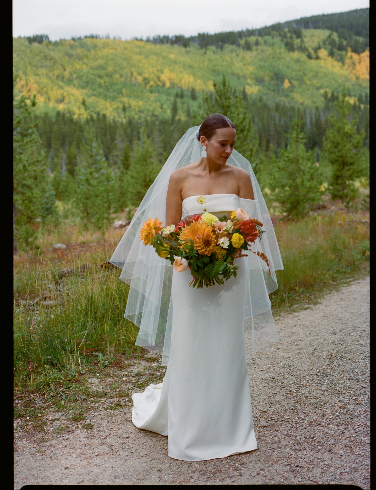 A Frame Club Fall wedding rocky mountain bride posing in nature with a large bouquet of autumnal flowers and a scenic mountain backdrop.