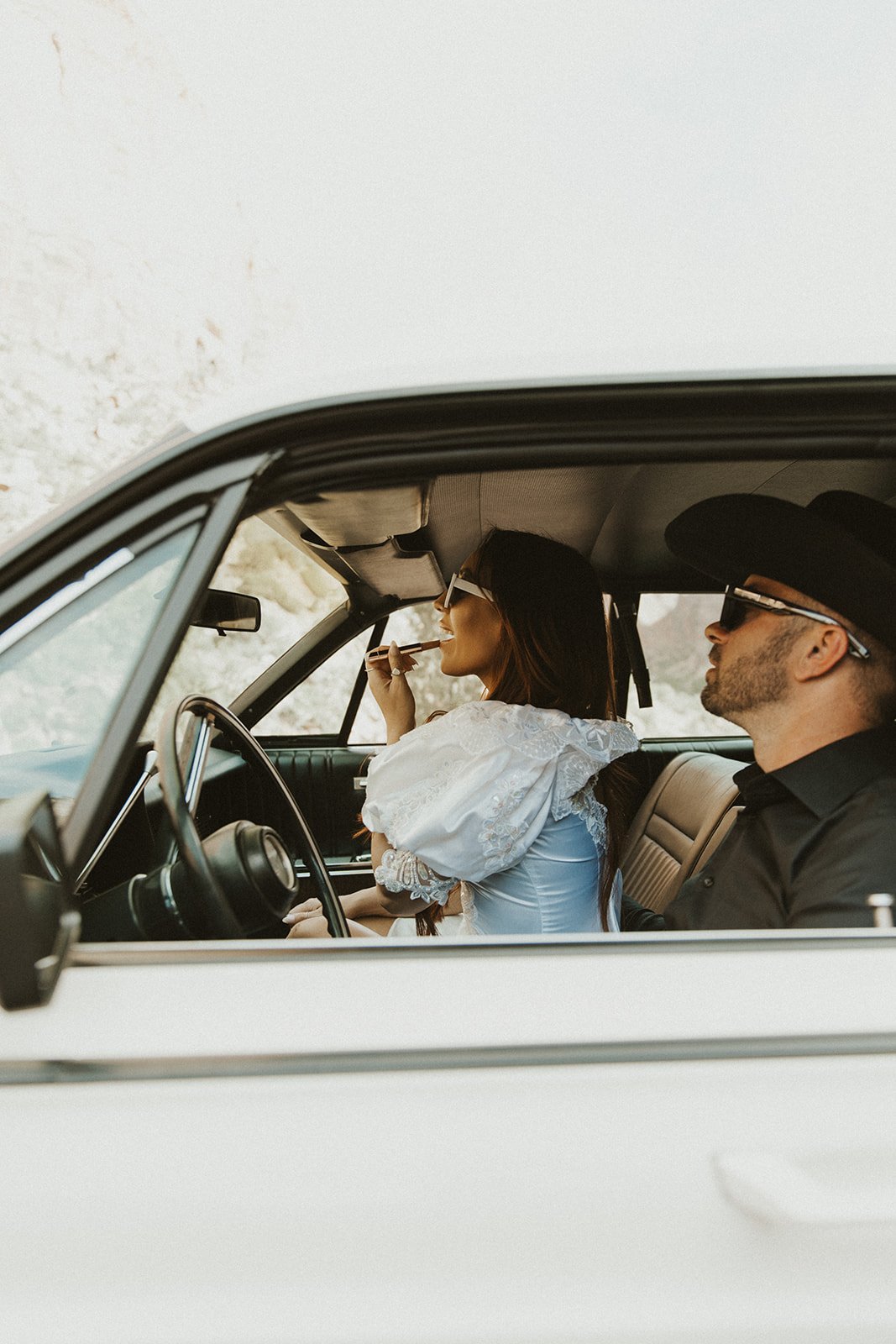 A bride in a puff-sleeve dress and groom in a black shirt share a stylish moment inside a classic white car surrounded by desert cliffs.