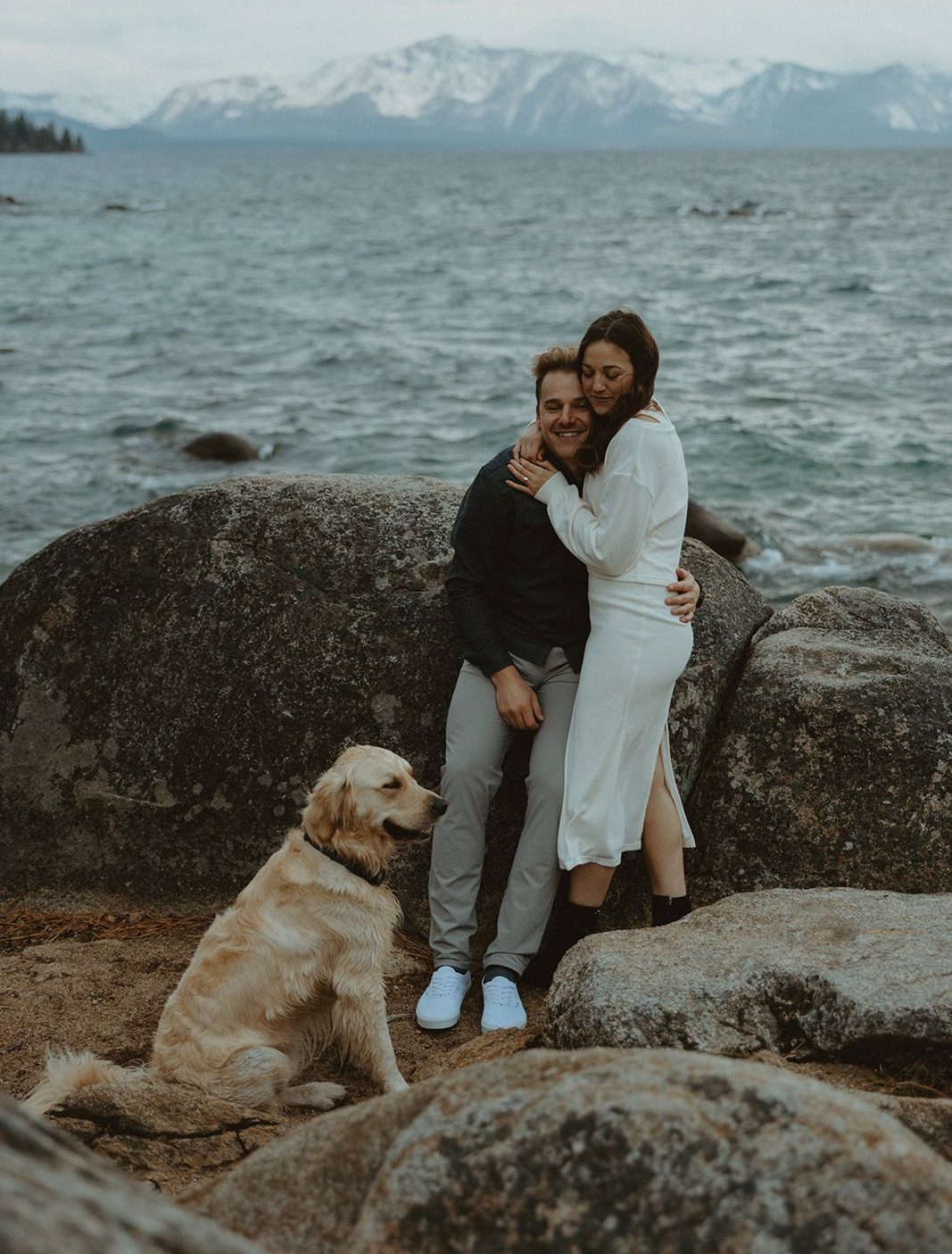 Couple cuddling on large rocks by the lake with their golden retriever during a cozy session captured by an Engagement Lake Tahoe Elopement Photographer.