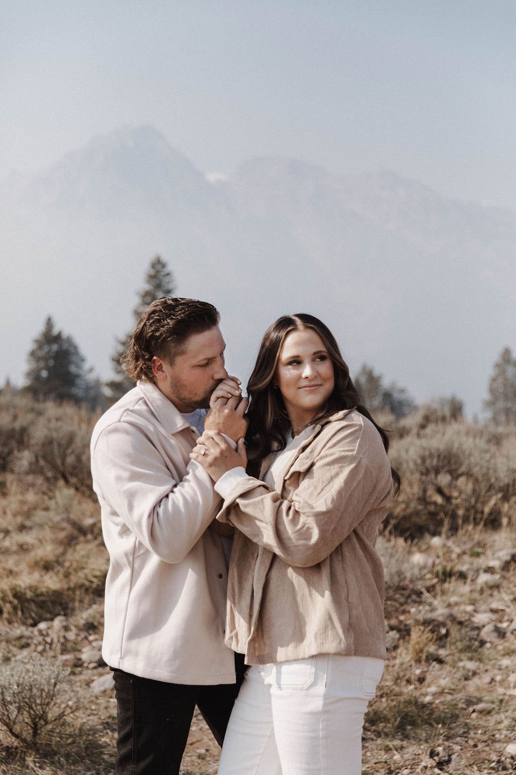 Tender kiss on the hand during a Grand Teton Engagement Session, with a couple dressed in neutral tones and stunning mountains in the distance.