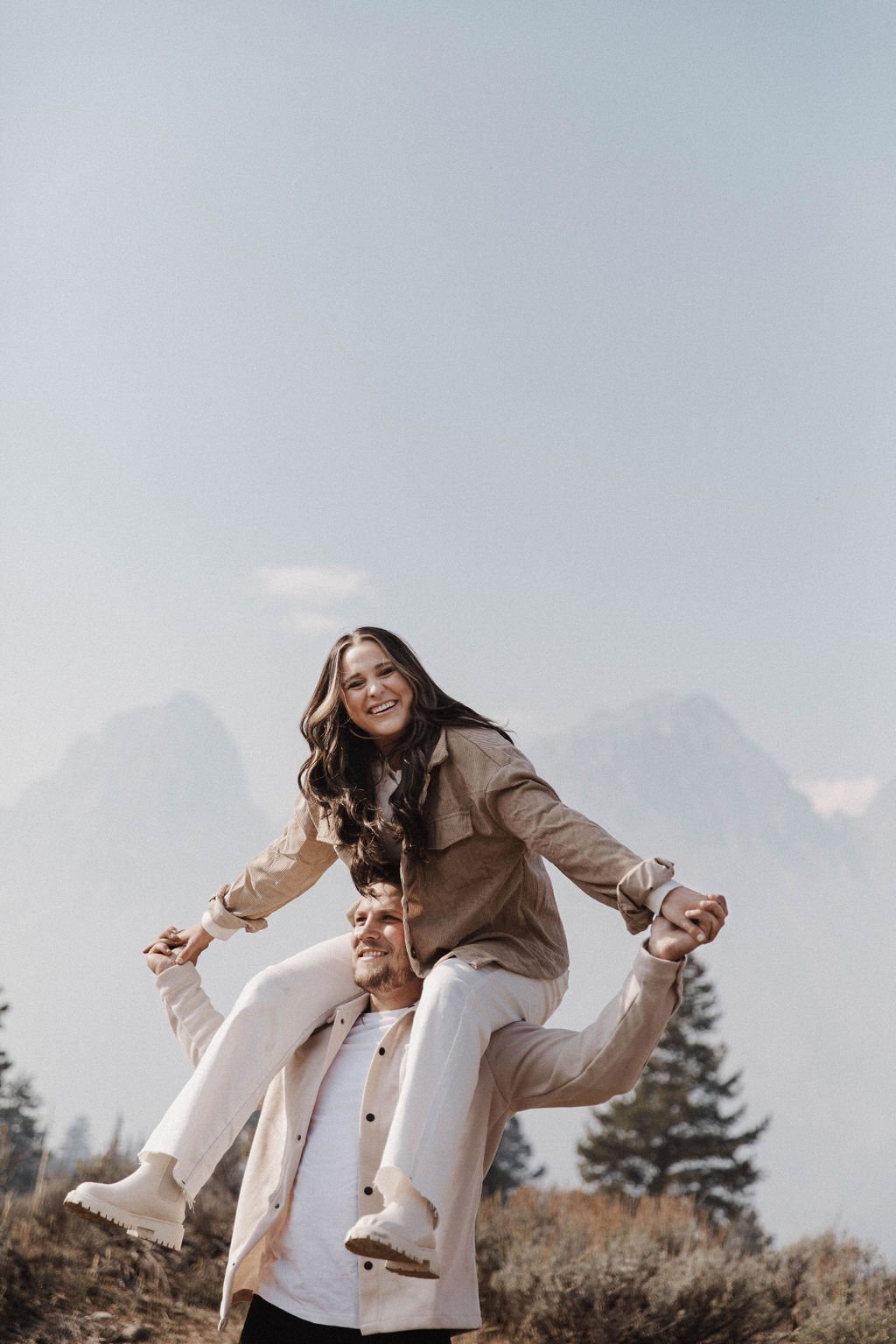 Joyful Grand Teton Engagement Session featuring a woman on her fiancé’s shoulders, smiling brightly with the mountains in the background.