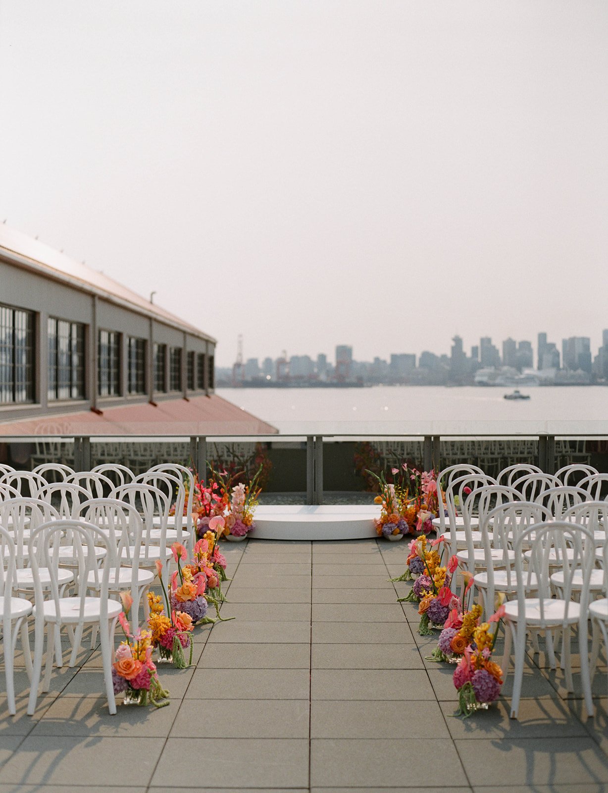 Elegant outdoor ceremony setup with white chairs and vibrant floral arrangements on a rooftop overlooking the city skyline at The Wallace Venue British Columbia wedding rocky mountain bride.