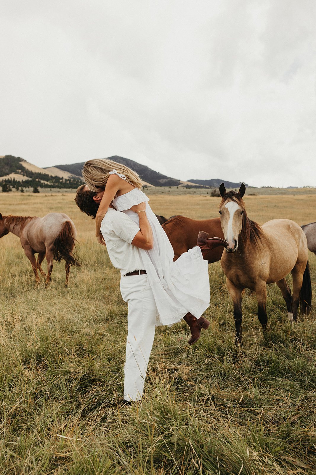 Man lifts his fiancée in a joyful embrace surrounded by horses in an open pasture during a Western summer engagement session in Montana.
