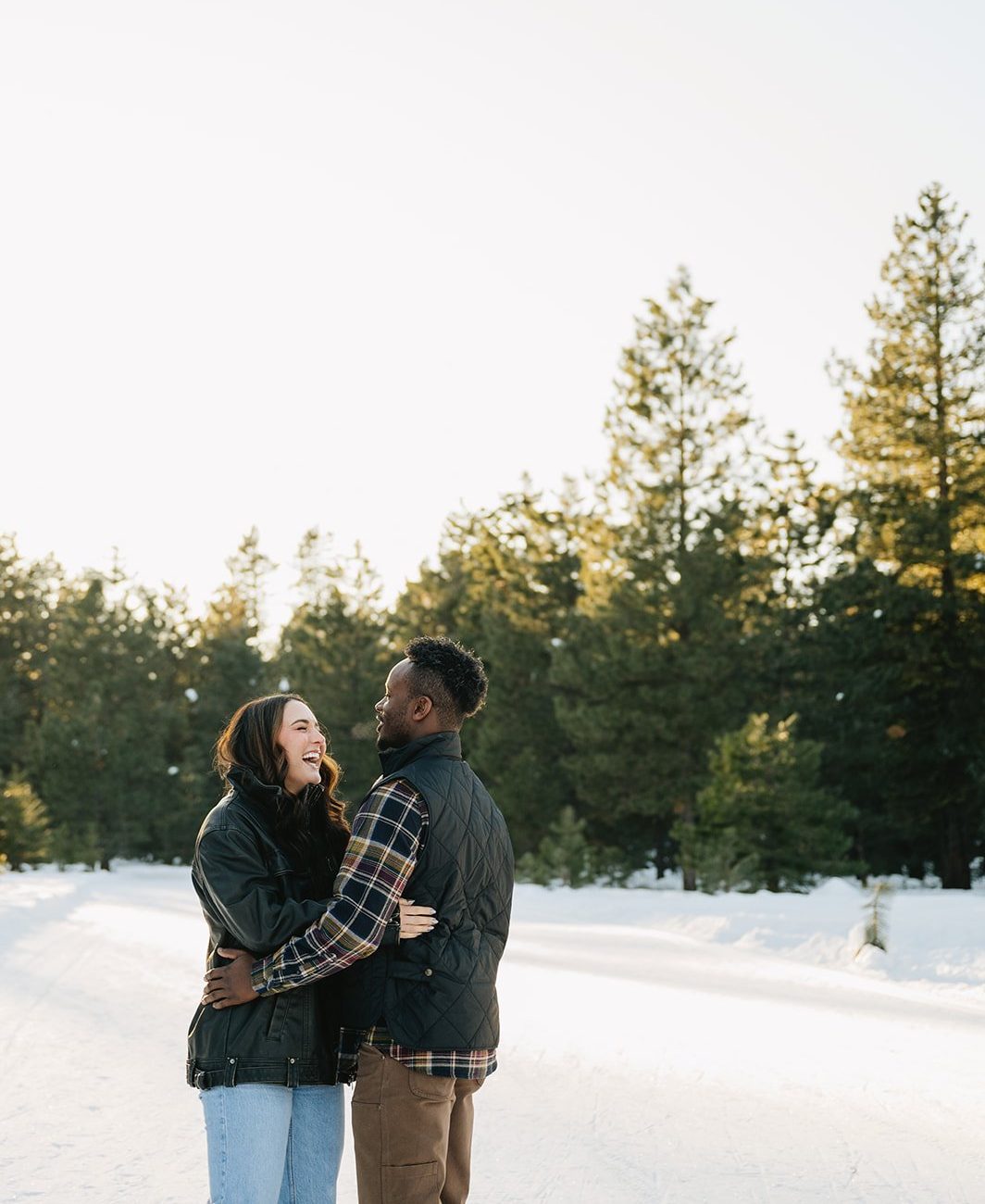 Engaged couple laughing together on a snowy forest path lined with pine trees during their Winter McCall Idaho Engagement Session.