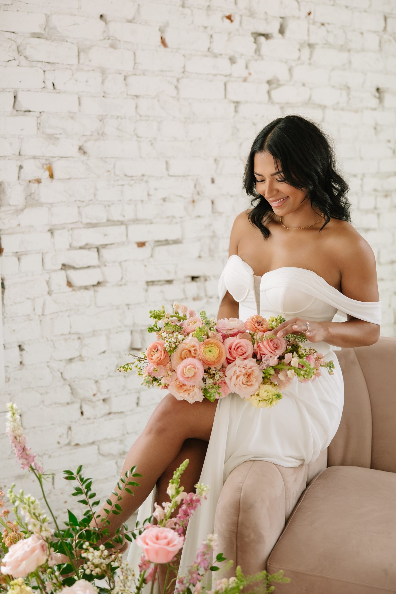Joyful bride holding a romantic peach-toned bouquet seated against a white brick wall for indoor utah wedding inspiration rocky mountain bride.