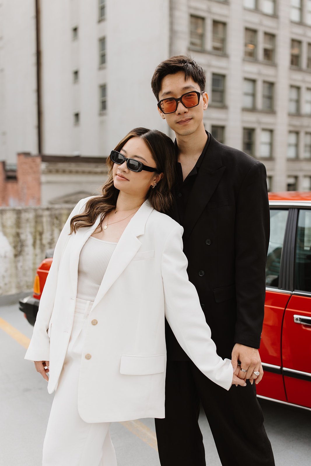 Close-up portrait of a stylish couple wearing black and white suits with sunglasses, leaning against a red car during their vancouver engagement miranda anderson photography shoot.