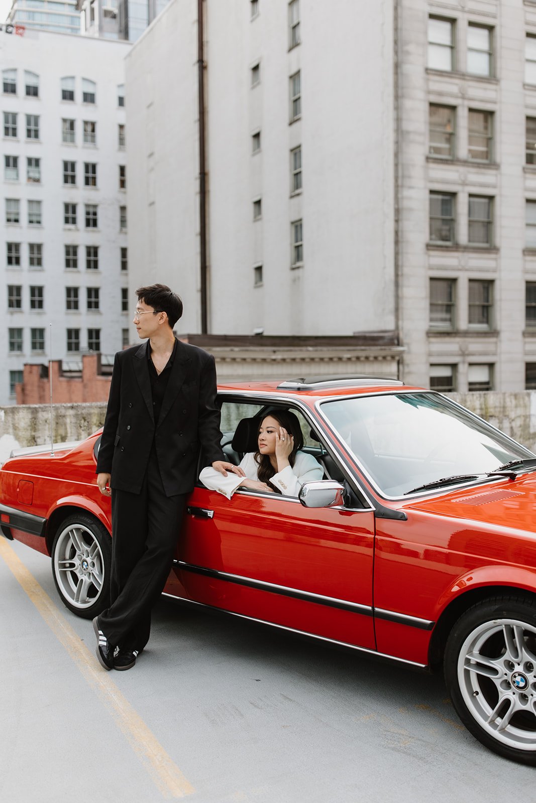 Couple poses beside a red vintage BMW on a rooftop with urban architecture in the background during a vancouver engagement miranda anderson photography session.