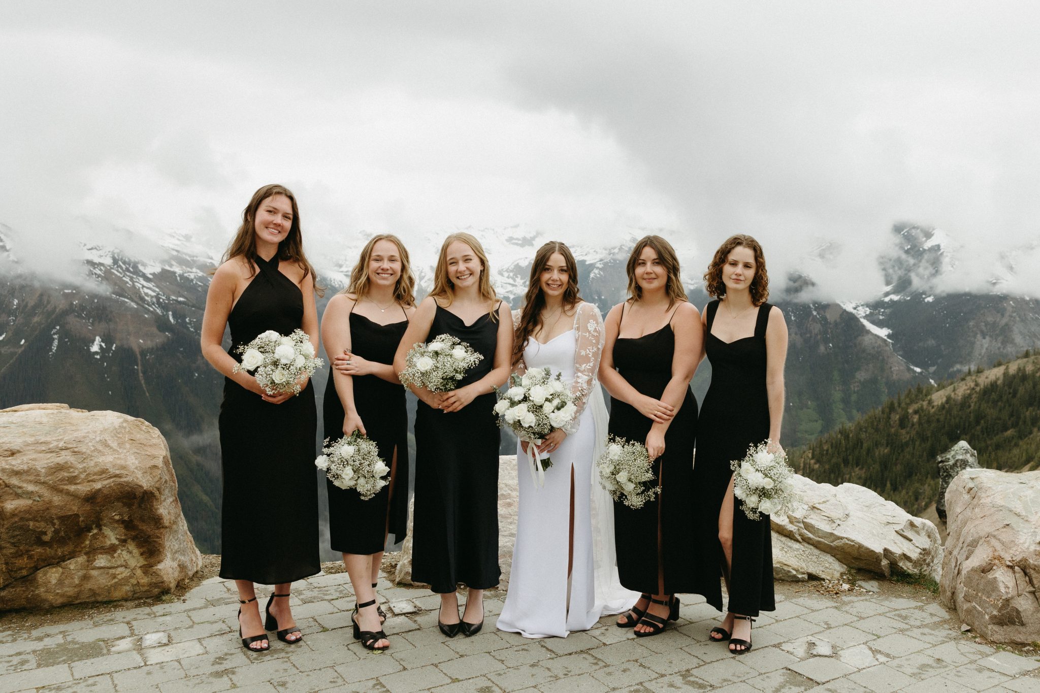 Bride poses with bridesmaids dressed in black at a scenic mountain overlook during a Golden british columbia spring wedding rocky mountain bride celebration.